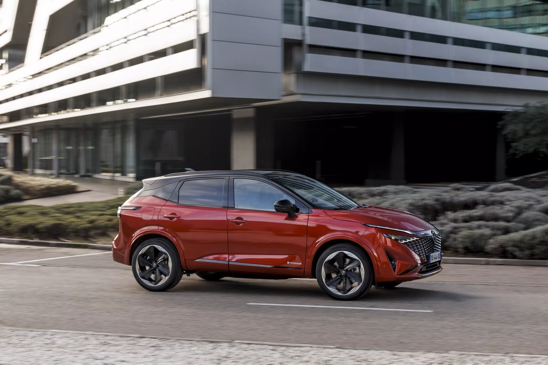 Dynamický boční profil červeného SUV Nissan Qashqai 2024 v pohybu, zdůrazňující jeho výšku a postoj z nízkého úhlu. Pohled ze tří čtvrtin ukazuje sportovní vícepaprsková litá kola, nízkoprofilové pneumatiky, tvarovanou aerodynamickou karoserii s výraznými rysy, ladná okna se zatmavením, zapuštěné kliky dveří, boční zrcátka s integrovanými směrovkami, pokročilé LED světlomety, výraznou přední mřížku, metalickou povrchovou úpravu a celkový odvážný současný městský design.