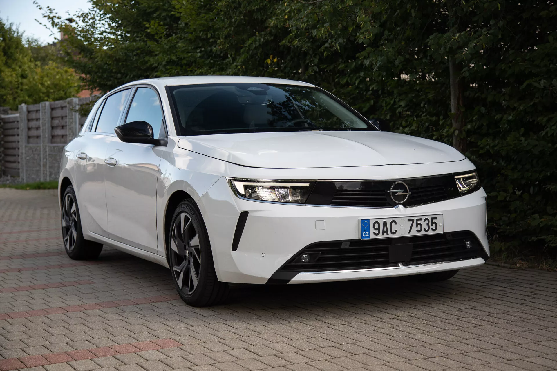 Opel Astra 2022 zachycený v předměstském prostředí, předvádějící své elegantní světlomety, výraznou přední mřížku, aerodynamický design včetně vícepaprskových slitinových kol a inovativní technologie z pohledu zepředu o tři čtvrtiny
