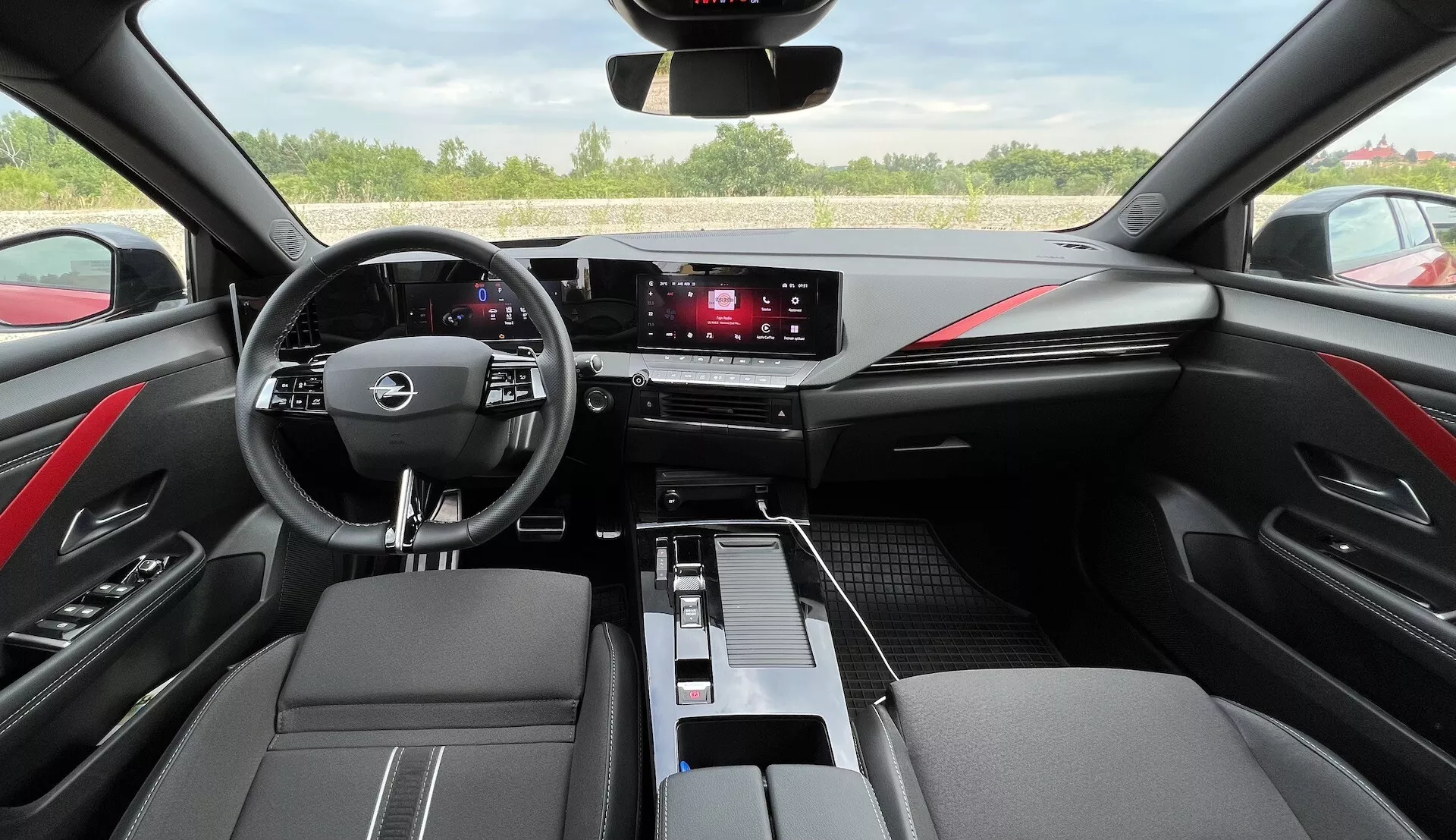 Interiér Opel Astra 2022 s multifunkčním volantem se třemi paprsky, digitálním přístrojovým panelem, dotykovým informačním systémem, elegantními ovladači klimatizace, černými kůží potaženými díly se červeným prošíváním a moderním designem kabiny z pohledu řidiče