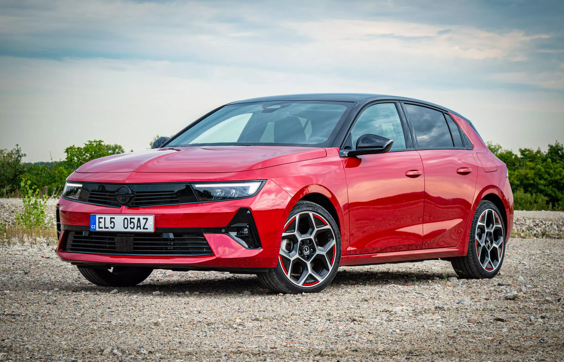 Červený hatchback Opel Astra 2022 fotografovaný z čelně-bočního pohledu ukazující LED světlomety, výraznou přední masku, kola z lehké slitiny s více paprsky a plynulý boční profil na štěrkovém povrchu s rozptýleným slunečním světlem osvětlujícím moderní prvky vozu