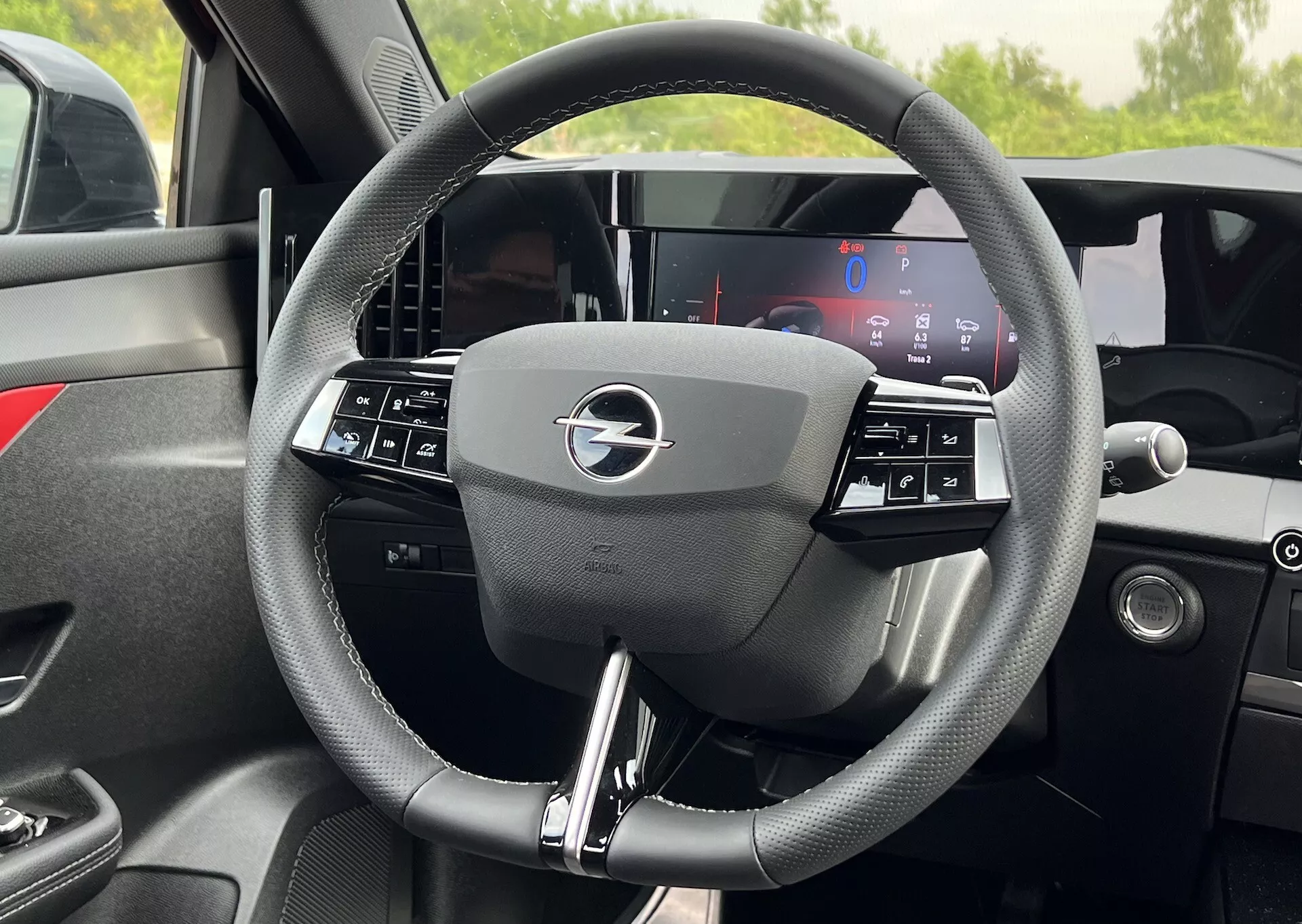 Detailní pohled zevnitř na volant Opel Astra 2022 s trojramenným designem a integrovanými ovládacími prvky a plně digitálním displejem palubní desky za ním, zdůrazňující moderní technologie a ergonomii vozidel
