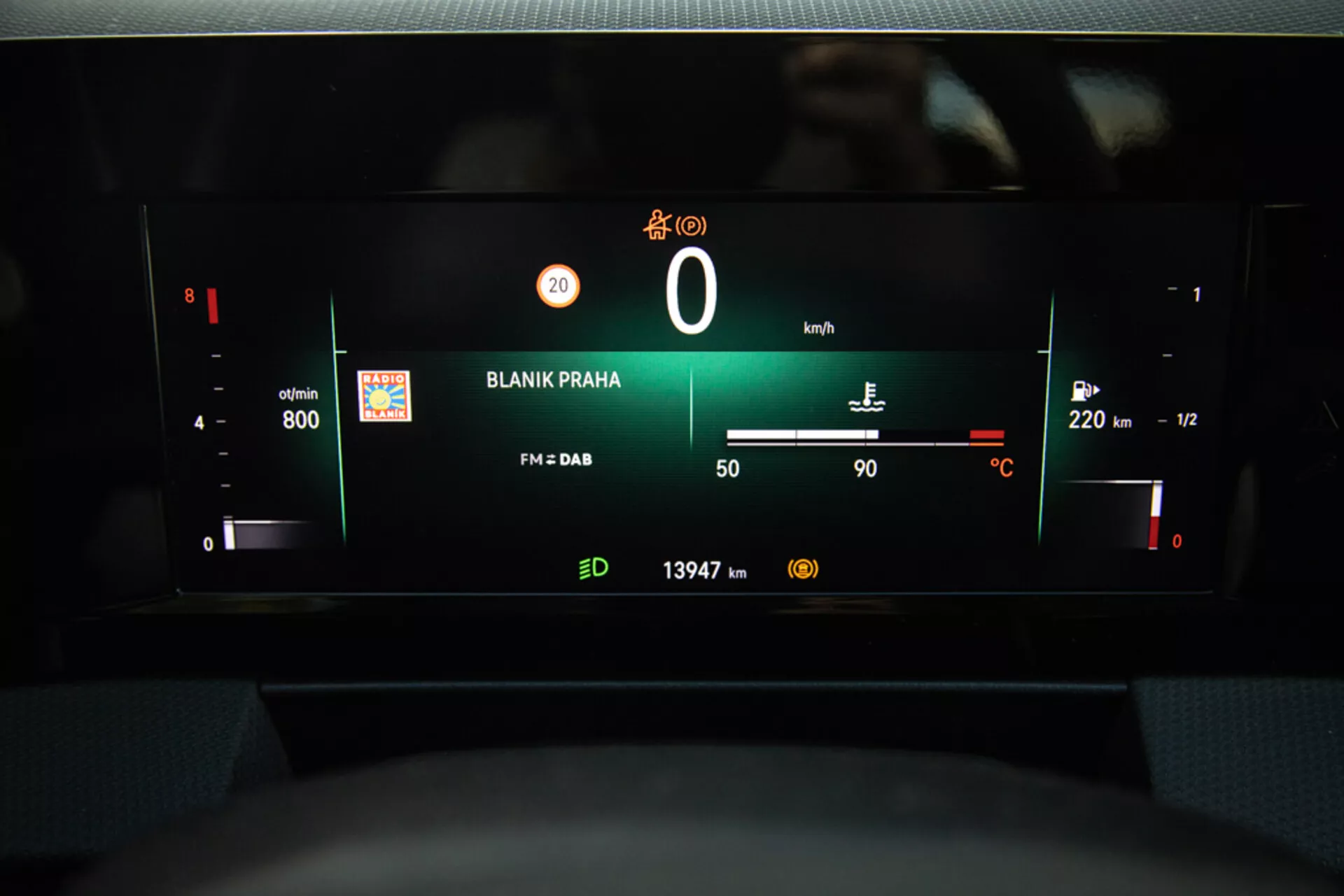 Pohled na digitální palubní desku Opel Astra 2022 s důrazem na přístrojový panel s otáčkoměrem, úrovní baterie, rychloměrem, ukazatelem paliva, vnější teplotou, rádiovou stanicí 'BLANIK PRAHA' a odečtem kilometrů z perspektivy řidičova sedadla.