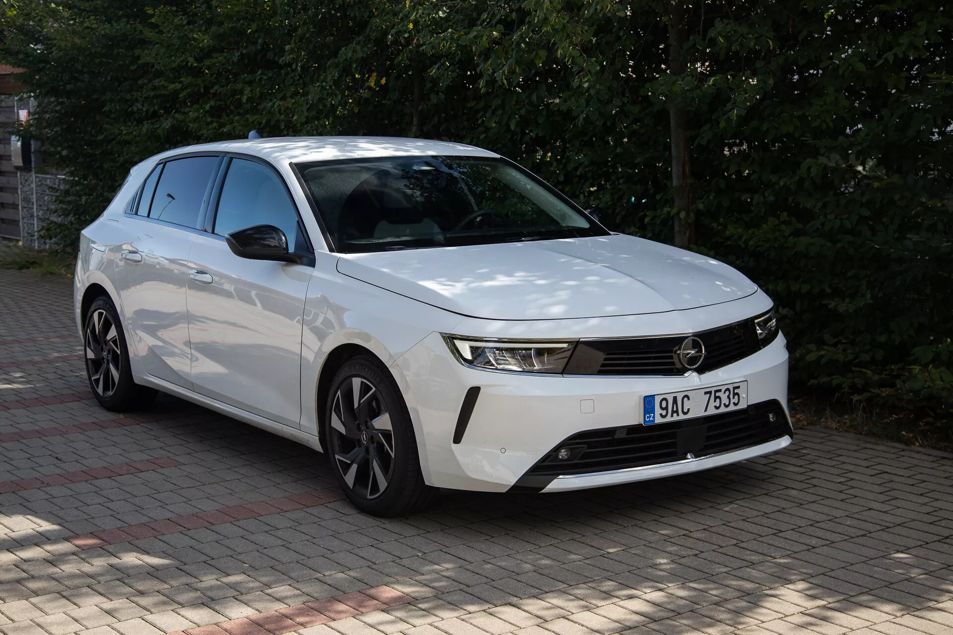 Bílá Opel Astra 2022 s výrazným designem přední části, štíhlými světlomety, horizontální mřížkou, aerodynamickým nárazníkem, profilovanou kapotou a ráfky z lehké slitiny zachycená z předního trojčtvrtěčního pohledu venku během dne