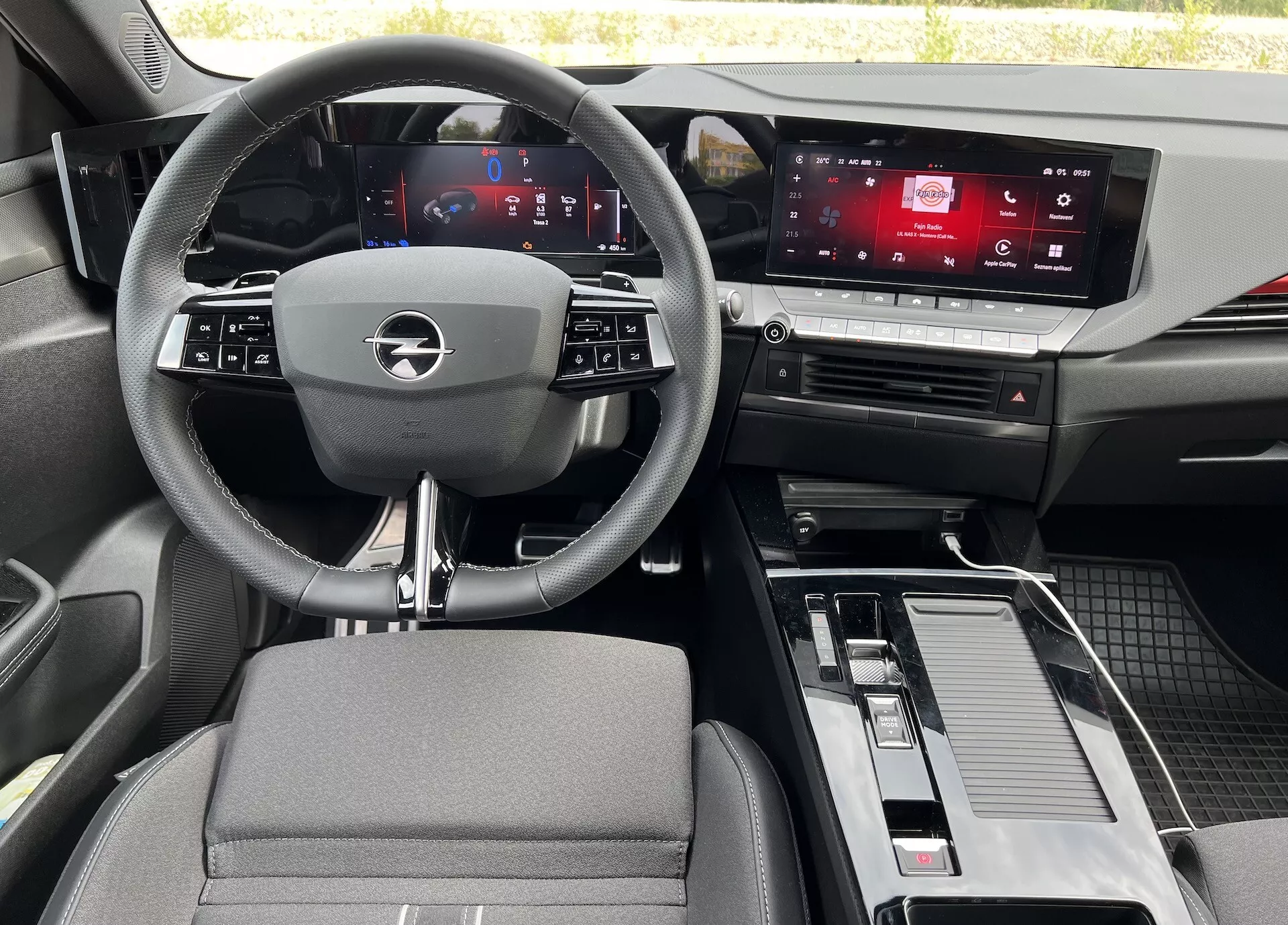 Interiér Opelu Astra 2022 z pohledu ze zadního sedadla, ukazující tříramenný volant s integrovanými ovladači, digitální přístrojový panel, dotykový displej infotainmentu, středovou konzolu s řadící pákou a úložným prostorem, a kombinaci materiálů z tkaniny a kovového efektu