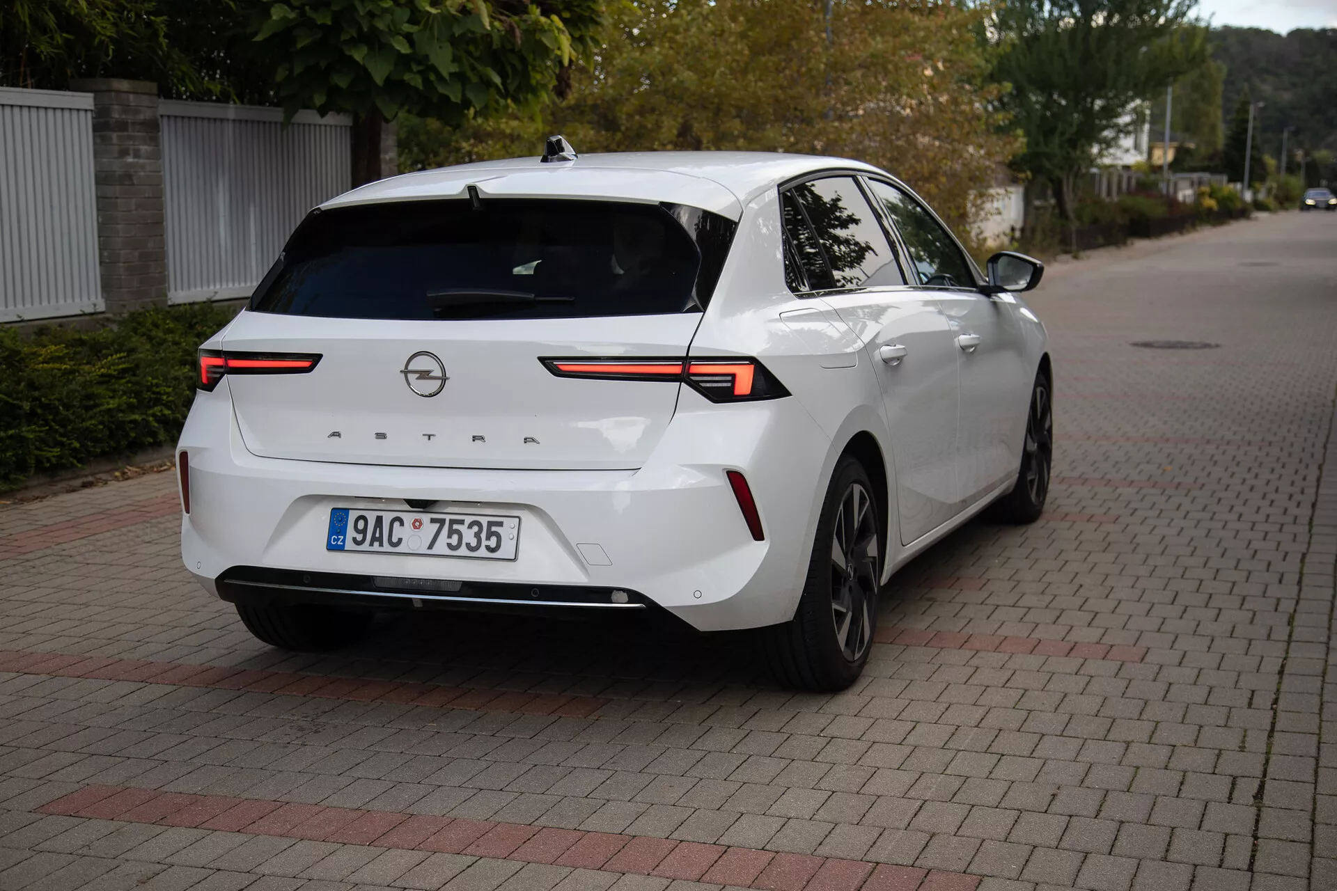 Moderní sedan Opel Astra 2022 v bílé barvě, zaparkovaný na povrchu ze zapadajících cihel, zobrazuje úhel tři čtvrtiny zadní části s důrazem na vyspělé LED zadní světla, zadní kameru, parkovací senzory, částečný boční profil včetně pravého zadního kola a dveří, spojený příčnou světelnou lištou přes víko zavazadlového prostoru, vybavený žraločí ploutví anténou pro zvýšenou konektivitu, vhodný pro ilustraci designu zadní části vozidla a moderní automobilové technologie