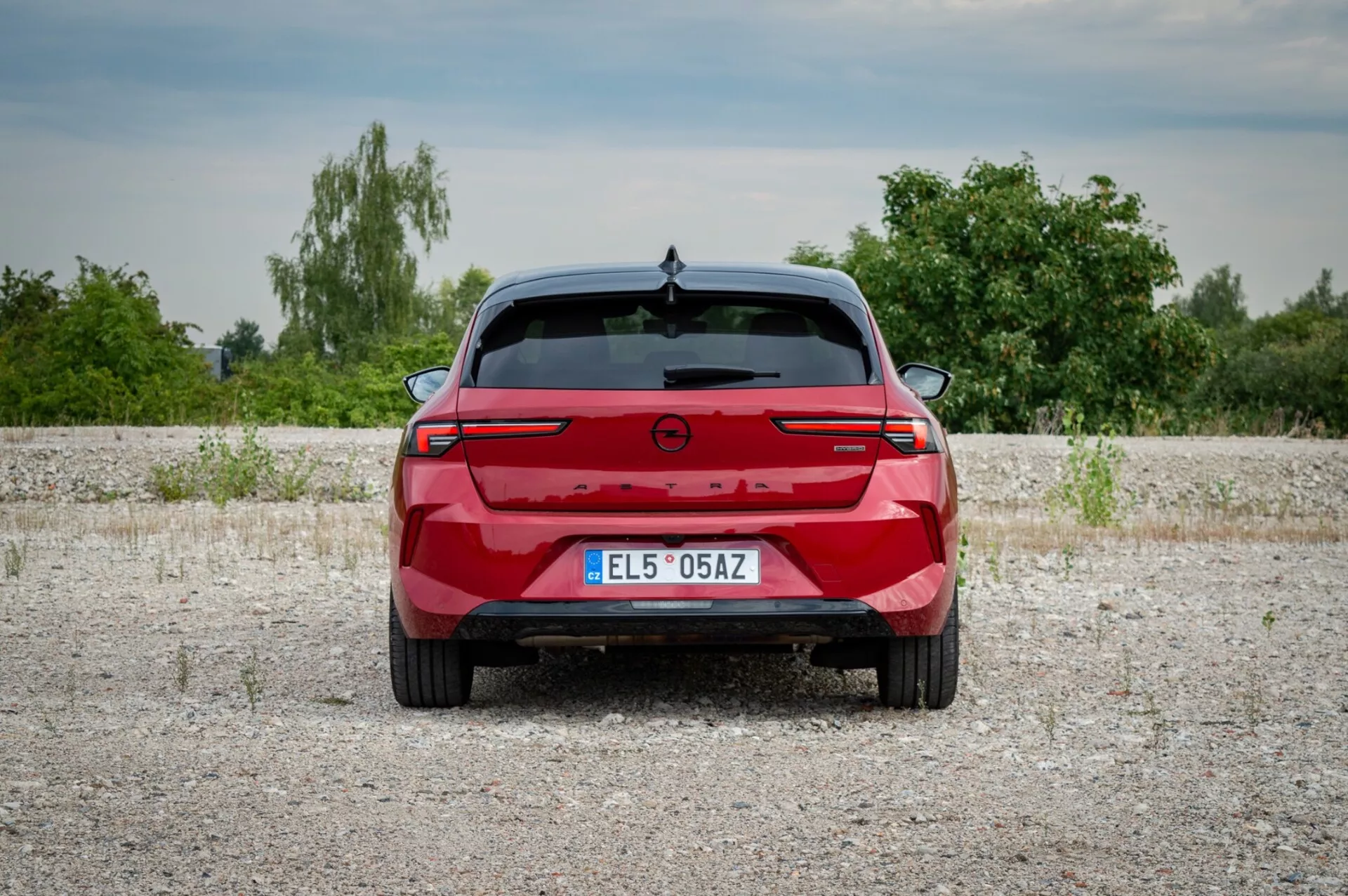 Opel Astra 2022 fotografovaný zezadu na štěrkovém povrchu s předvedením zadních světel s propojovací světelnou lištou, parkovacími senzory a absencí výfukových portů naznačující možný elektrický nebo hybridní model, na pozadí vegetace a mírně zamračené oblohy.
