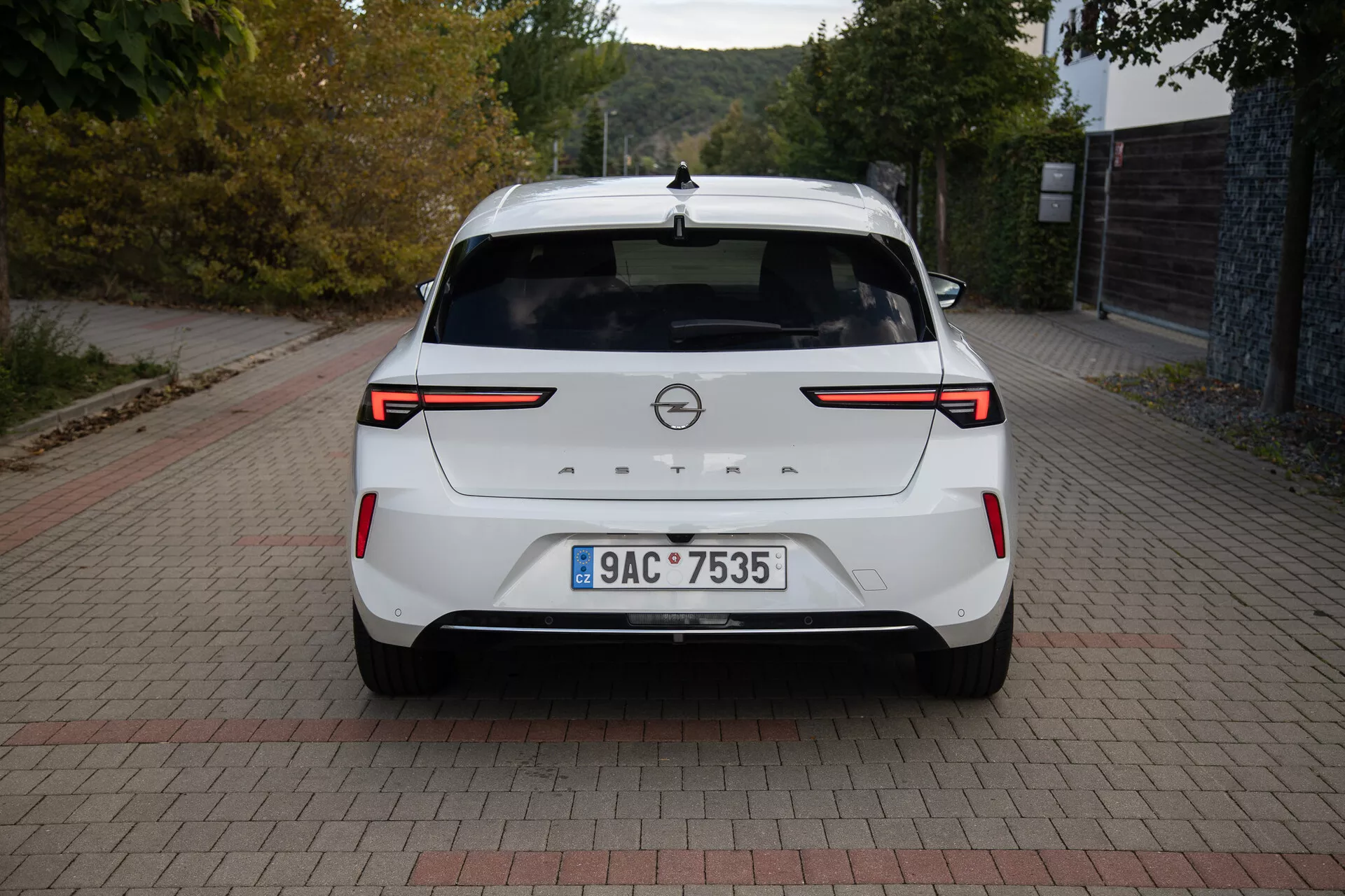 Zadní pohled na bílý Opel Astra 2022 s LED zadními světly, parkovacími senzory / kamerou vzadu, znakem značky, registrační značkou, zadním spoilerem, vysoko umístěnou stop svítilnou, anténou ve tvaru žraločí ploutve a čistým lakem, zaparkovaný na cihlově vzorovaném povrchu ve dne.