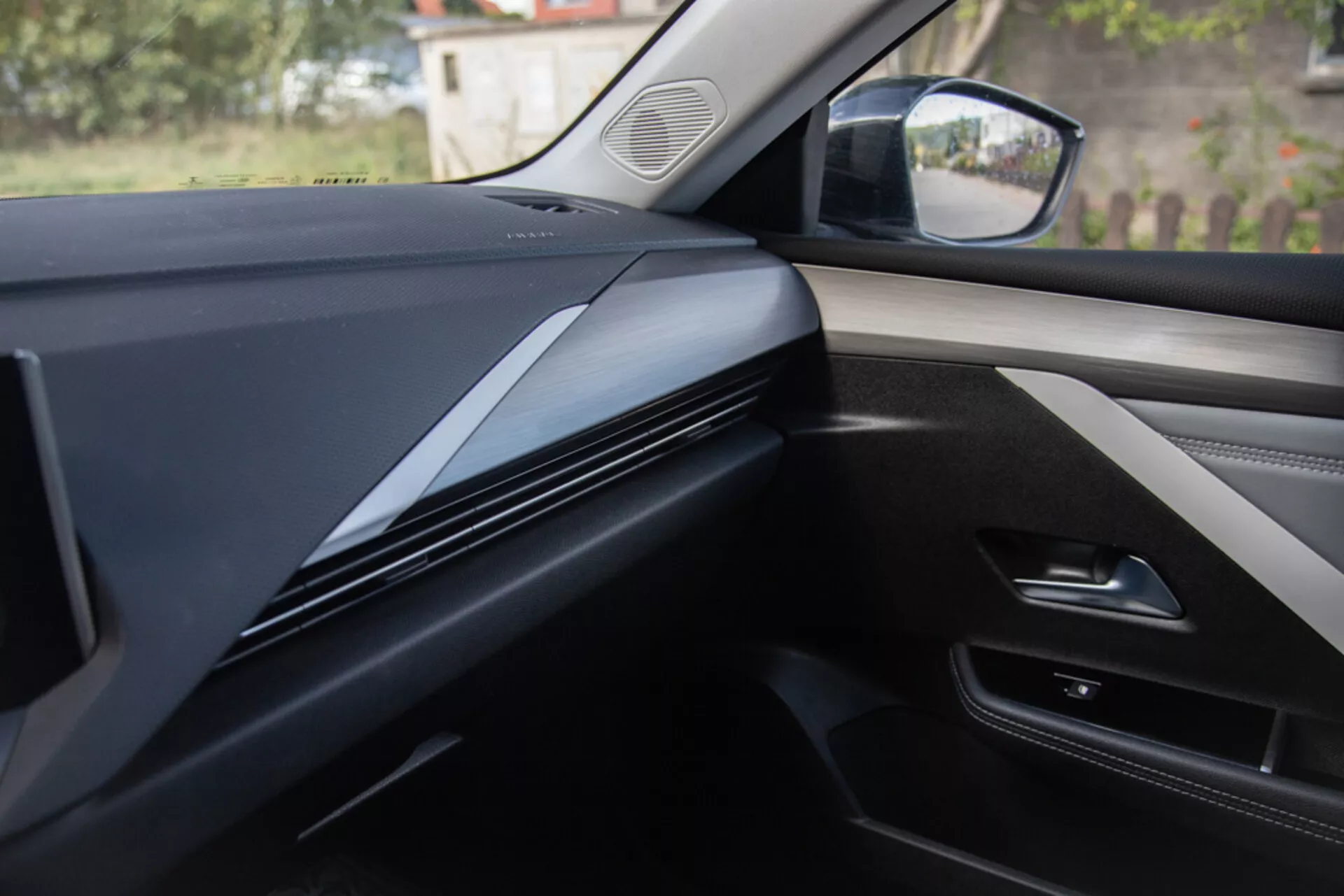 Vnitřní pohled z perspektivy spolujezdce ukazuje design palubní desky, ozdobné lišty a dveřní panel v Opelu Astra 2022, s tmavými, elegantními konturovanými liniemi se zaměřením na texturu materiálu a potenciální technologické integrace