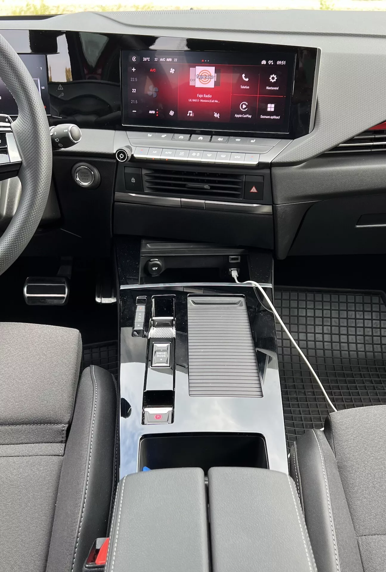Interiér Opelu Astra 2022 ukazující moderní design kabiny s dotykovým informačním systémem, dotykovými ovladači klimatizace, kovovou řadicí pákou, USB konektivitou, držáky na nápoje, detailně zpracovanou středovou opěrkou, sedadly ve dvoubarevném provedení a detaily kabiny z kombinace materiálů s odolnými podlahovými rohožemi