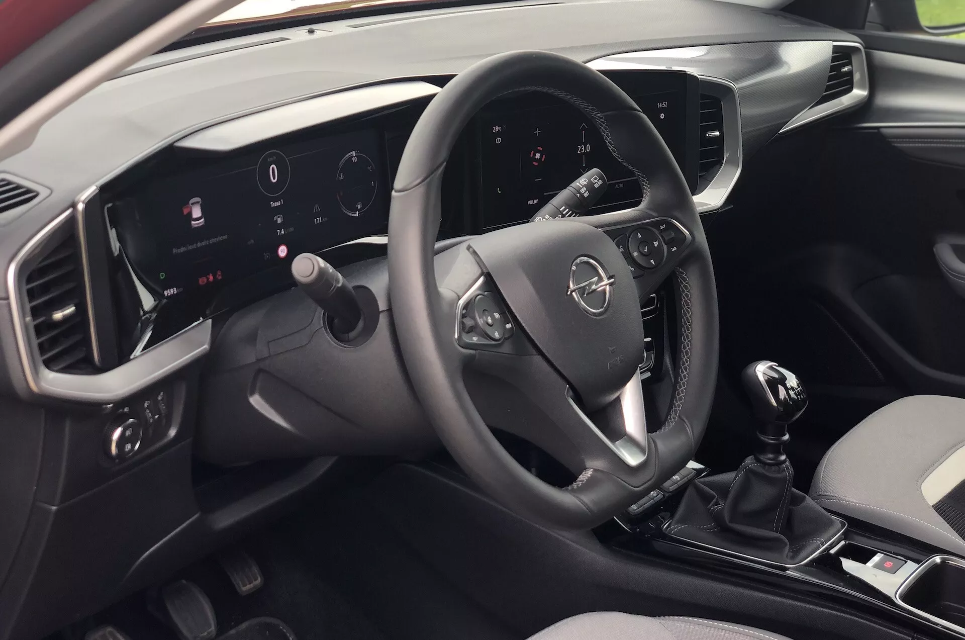 Vnitřní pohled na Opel Mokka 2021 z perspektivy sedícího na zadním sedadle napravo, zobrazující moderní palubní desku, multifunkční volant, osvětlený digitální palubní displej, dotykový infotainment systém, manuální řazení a elegantní dvoubarevný design.