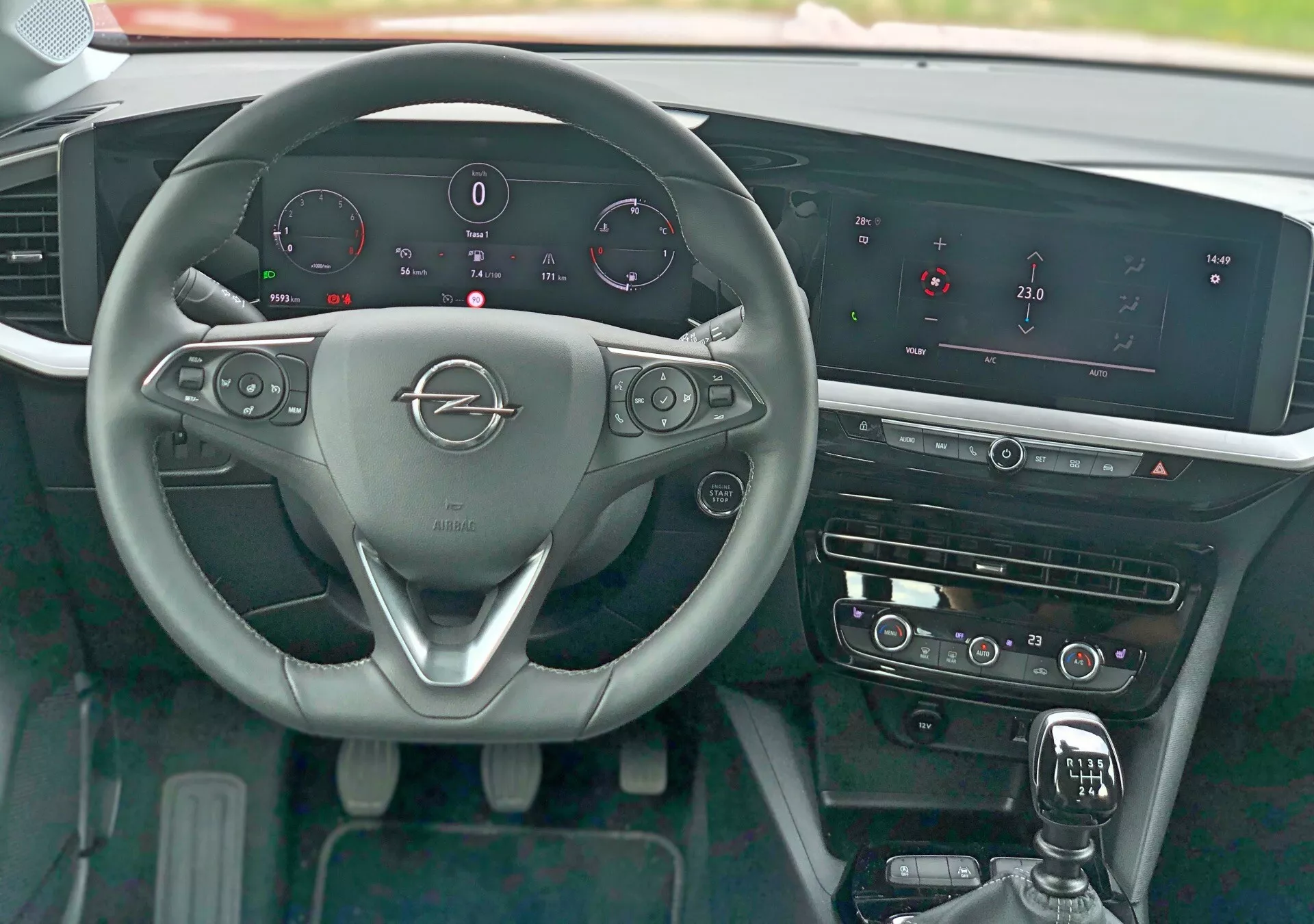 Pohled z interiéru Opel Mokka 2021 z pohledu řidiče ukazuje tříramenný volant s kovovým logem značky, ovládací prvky volantu pro audio, telefon a tempomat, pádla pro řazení, digitální palubní desku zobrazující rychlost, otáčky, hladinu paliva a teplotu motoru, výrazný infotainment systém se širokou horizontální obrazovkou, dotykové ovládání HVAC, řadící páku s koženým převlečníkem, leštěné ozdobné prvky a tlačítko bezklíčového startování.