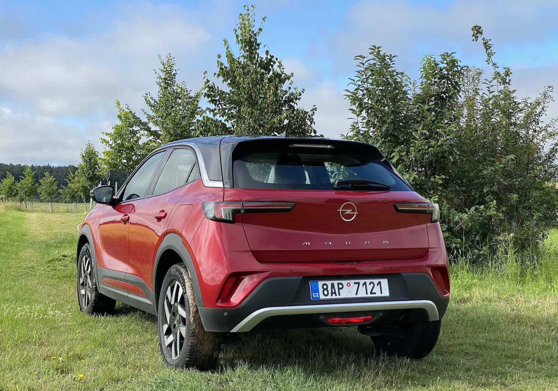 Barevná fotografie zobrazující zadní tři čtvrtiny moderního crossover SUV Opel Mokka 2021 v červené a černé barvě, zaparkovaného venku na travnaté ploše s keři nebo stromy v pozadí. Designové prvky vozidla zahrnují aerodynamický zadní spoiler, hranaté LED zadní světla, vysoko umístěnou brzdovou lampu, dvoubarevné tělo, velká litá kola, zapuštěné kliky dveří a černé plastové opláštění. Fotografie je pořízena z nízkého úhlu, což zdůrazňuje výšku a světlou výšku SUV, a obloha je částečně zatažená, což naznačuje proměnlivé počasí.