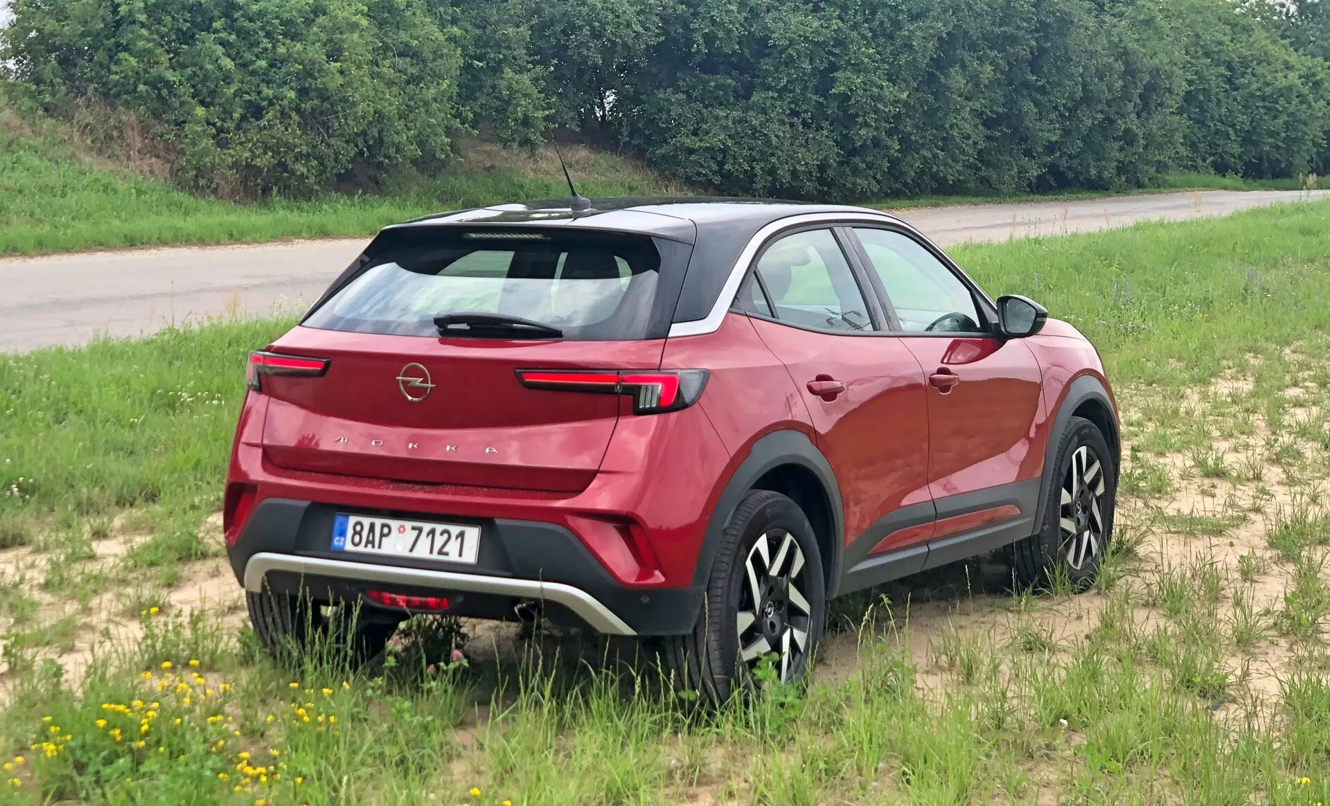Moderní crossover SUV Opel Mokka 2021 z pohledu ze zadní tří čtvrtin, zdůrazňující zadní spoiler, ostrý zadní sklo, viditelný znak značky a elegantní zadní světla. Vozidlo je umístěné na travnatém povrchu, což ukazuje jeho lehkou terénní potenciál. Boční profil ukazuje tvarované linie karoserie, černé obklady a stylová vícepaprsková kola. Integrovaný zadní stírač, parkovací senzory a kontrastní kryt podvozku naznačují aerodynamiku, praktičnost a robustnost.
