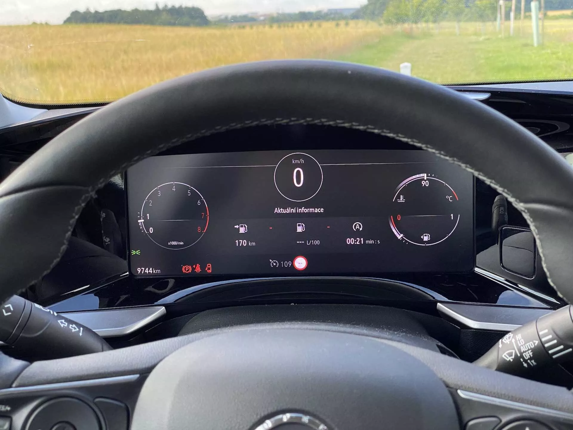 Interiérový pohled na Opel Mokka 1.2 DI Turbo 2021 z pohledu řidiče za volantem. Obrázek zdůrazňuje digitální přístrojovou desku za volantem, která zahrnuje rychloměr, otáčkoměr a informační panel zobrazující venkovní teplotu (8°C), hodiny, polohu převodovky ('P' pro parkování) a podrobnosti palubního počítače. Volant obsahuje viditelné ovládací prvky pro média a je částečně viditelný. Přirozené osvětlení naznačuje denní dobu a přes čelní sklo je vidět venkovní výhled na pole. Vozidlo ukazuje elektronickou zobrazovací palubní desku a pokročilé technologické funkce.