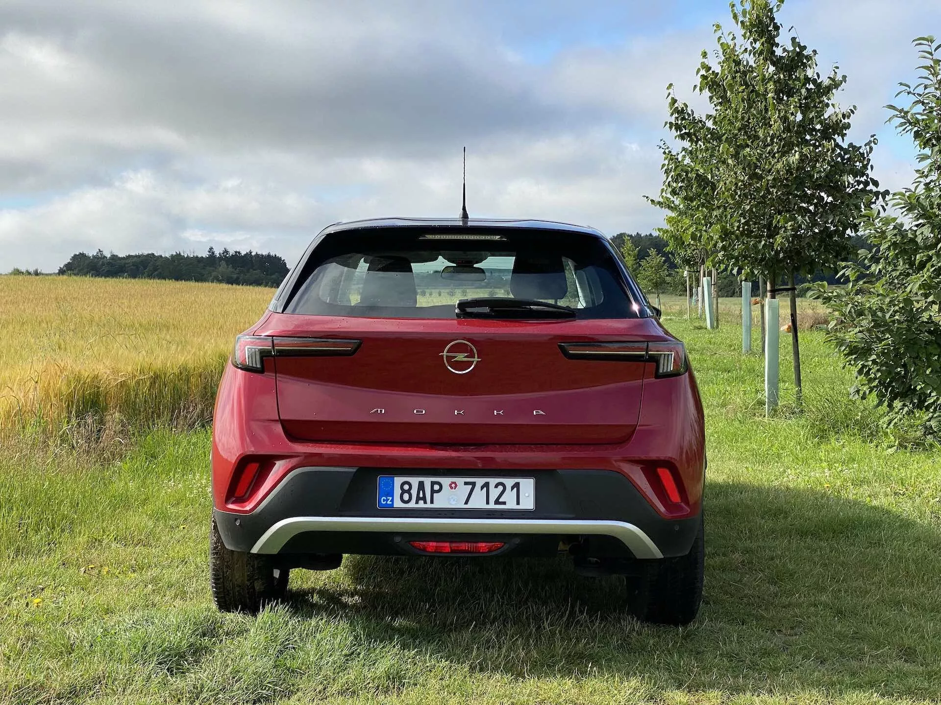 Zadní pohled na červený Opel Mokka 1.2 DI Turbo 2021, s moderním designem ostrých linií a emblémem výrobce na víku zavazadlového prostoru, výraznými LED zadními svítilnami, zadním nárazníkem s kontrastními vsadkami, parkovacími senzory, integrovanými odrazkami, stěračem zadního skla, žraločí anténou, zadním spoilerem a registrační značkou ve venkovském prostředí s trávníkem, polem a stromy.
