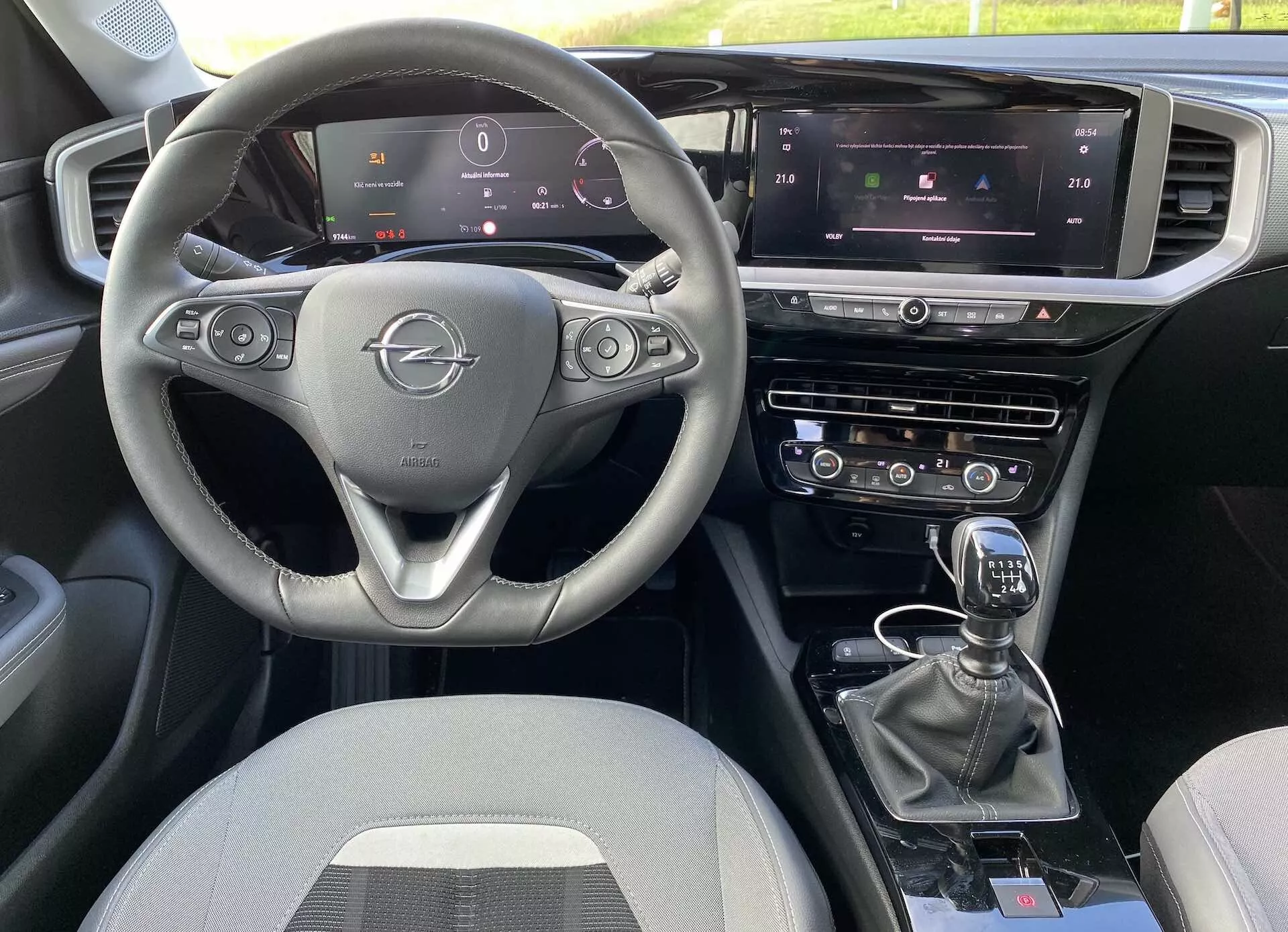 Interiér Opel Mokka 2021 z perspektivy předního spolujezdce, tříramenný volant s ovládacími prvky, plně digitální přístrojový panel, centrální infotainmentový dotykový displej s ovládáním klimatizace a navigace, a manuální řadicí páka. Rozložení zahrnuje duální řídicí systémy a další tlačítka pro jízdní režimy, všechno s látkovým čalouněním.