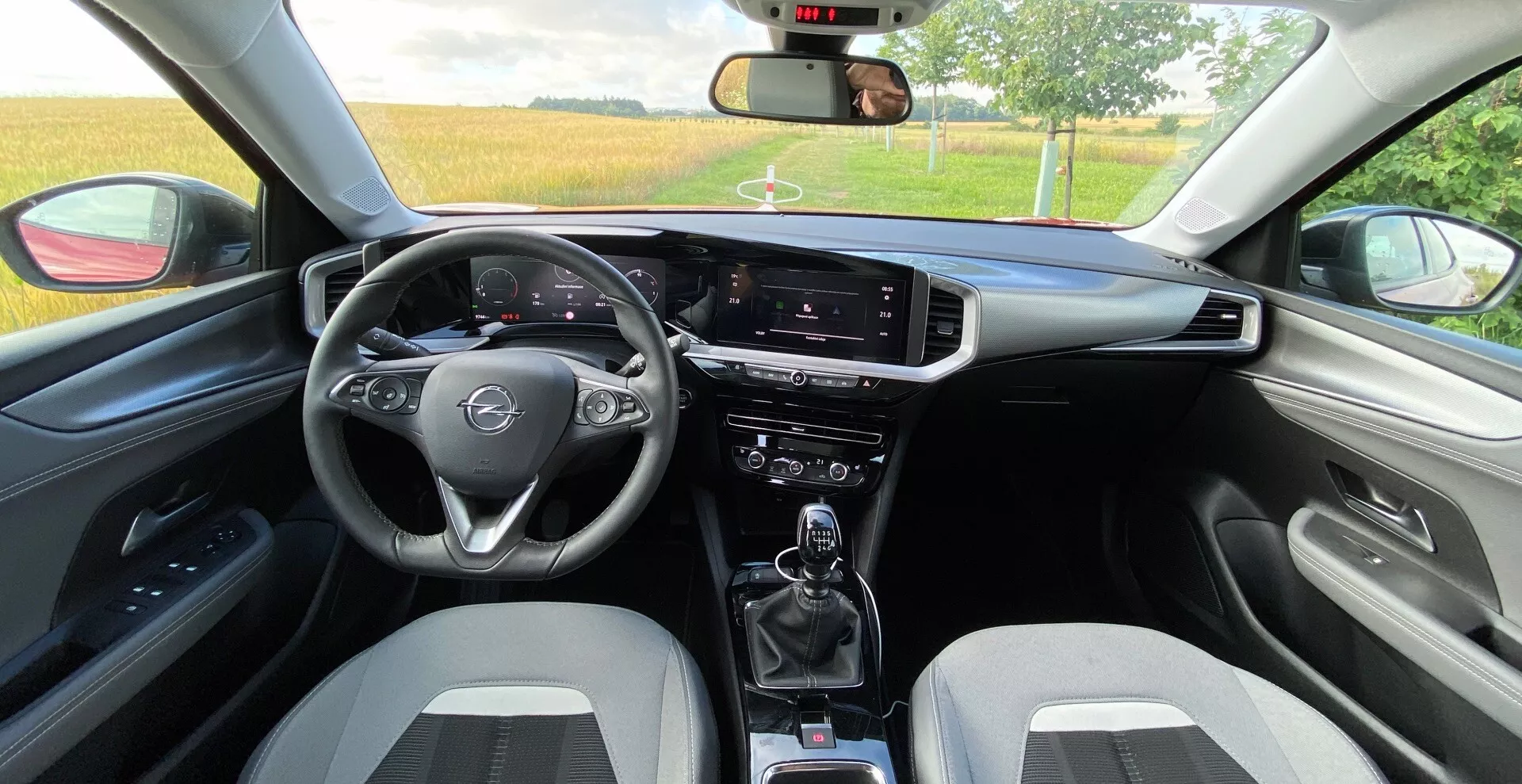 Pohled na interiér zezadu sedadla u Opel Mokka 2021 1.2 DI Turbo. Obsahuje tříramenný multifunkční volant, digitální rychloměr a otáčkoměr za volantem, infotainmentový dotykový displej na výšku, přepínače klimatizace, manuální převodovku s držáky nápojů, dvoubarevná sportovní přední sedadla, sladěné dveřní panely s područkou, viditelné zpětné zrcátko, částečně otevřená okna a další ovládací páčky pro stěrače a ukazatele směru.