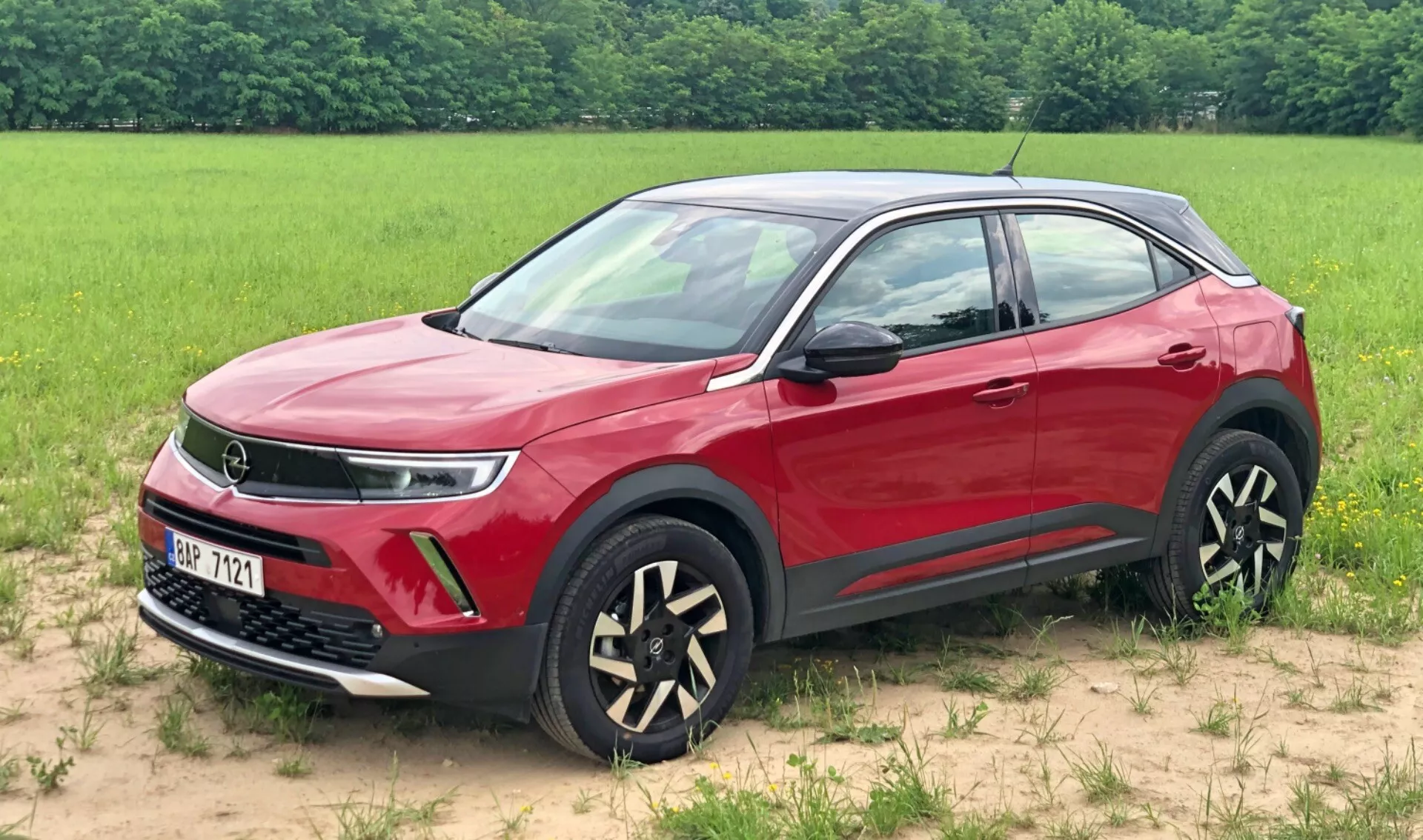Obrázek červeného vozu Opel Mokka 2021 SUV zaparkovaného na travnatém povrchu s polem v pozadí. Tento tříčtvrtinový pohled zepředu zdůrazňuje mřížku, potenciálně naznačující pokročilou pohonnou technologii, elegantní LED světla pro denní svícení, mlhovky a jedinečná dvoubarevná kola z lehkých slitin. Viditelná jsou také barevně slazená boční zrcátka a kliky dveří, dynamická linie ramen, ochranný černý spodní lem a chromové rámy oken, které se vyznačují moderními, luxusními designovými prvky vozu.