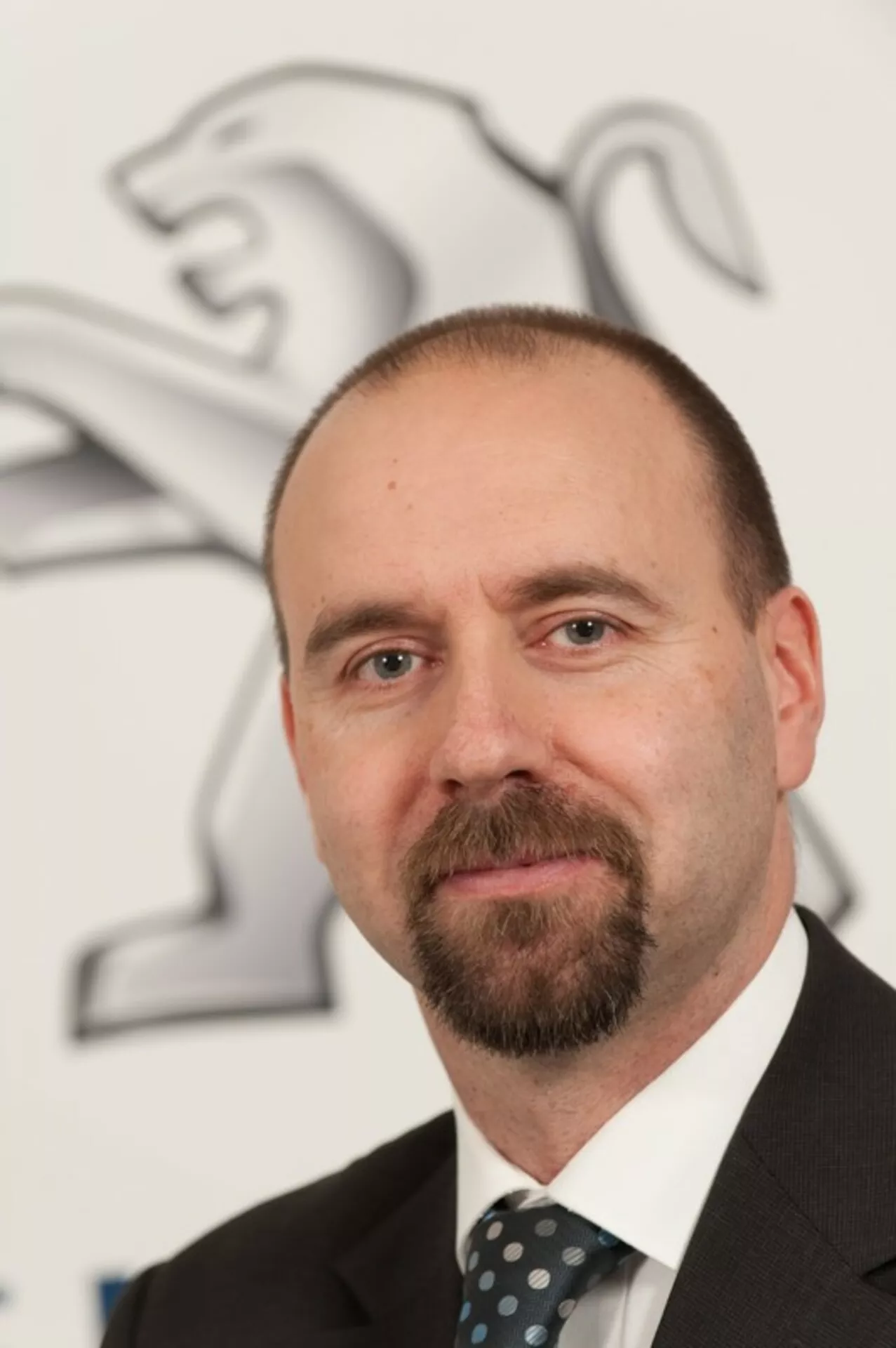 Profesionální portrét muže v obleku a kravatě s logem Peugeotu s lvem na pozadí, auto není na obrázku vidět