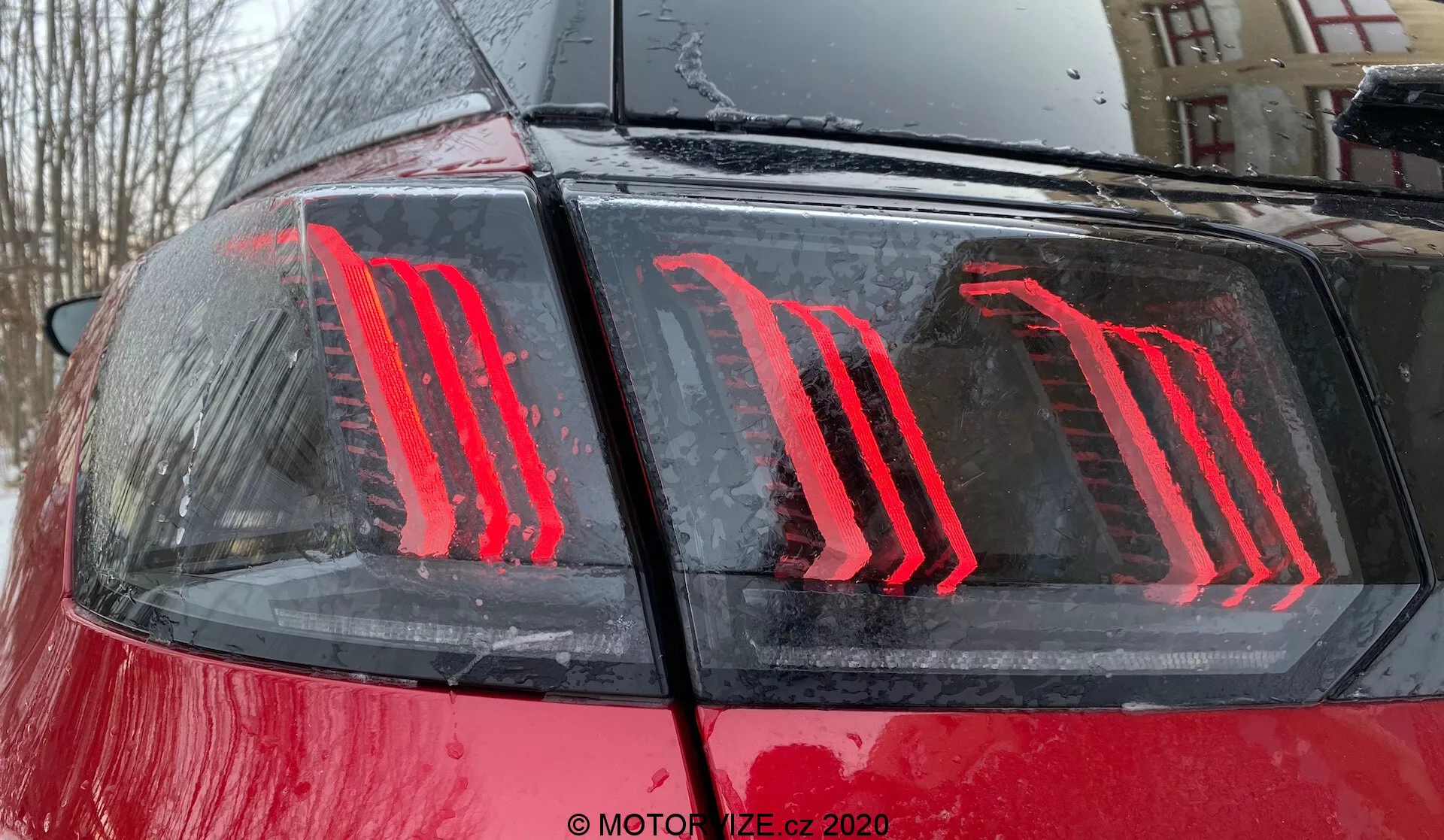 Detailní záběr na zadní část vozu Peugeot 3008 z roku 2021, zdůrazňující moderní svislé LED zadní světlo na lesklém červeném laku s kapkami vody, což naznačuje venkovní prostředí během nebo po dešti; zadní světlo obsahuje energeticky účinnou technologii a slouží jako brzdové a denní světlo.