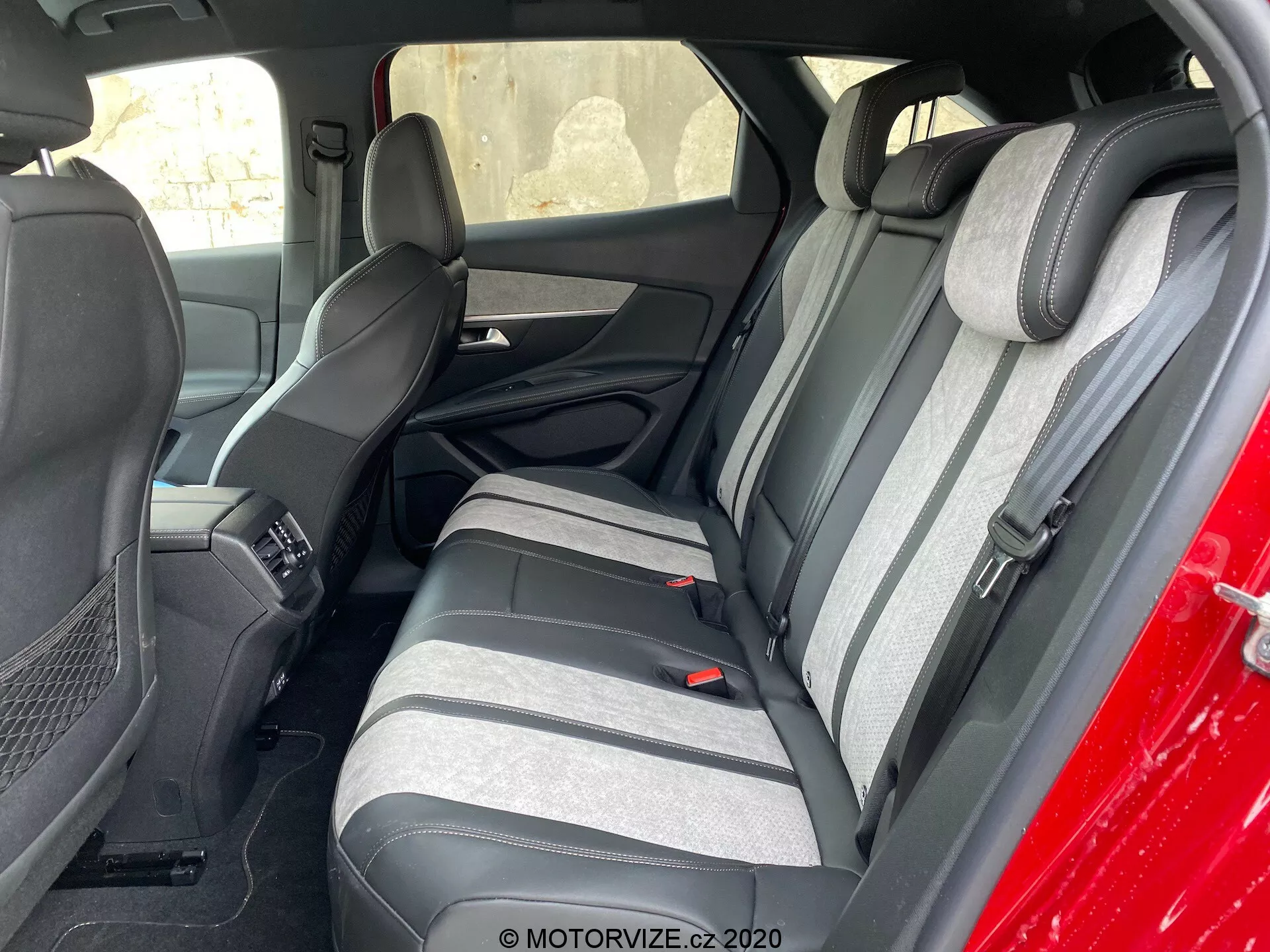 Snímek interiéru vozu Peugeot 3008 z roku 2021 zaměřený na zadní sedadlovou část, viděný zvenčí, šikmo k řidičově straně. Zadní sedadla jsou potažena dvoubarevnou látkou v tmavých a světlých odstínech šedé s texturovaným proužkem. Vidět jsou také odpovídající vyvýšené opěrky hlavy. Část předních sedadel a středová konzola jsou rovněž viditelné. Zadní sedadla mají tříbodové bezpečnostní pásy a opěrku s integrovanými držáky na pohárky. Interiérové panely dveří ladí s barvami sedadel a mají mřížky reproduktorů a ovládací prvky.
