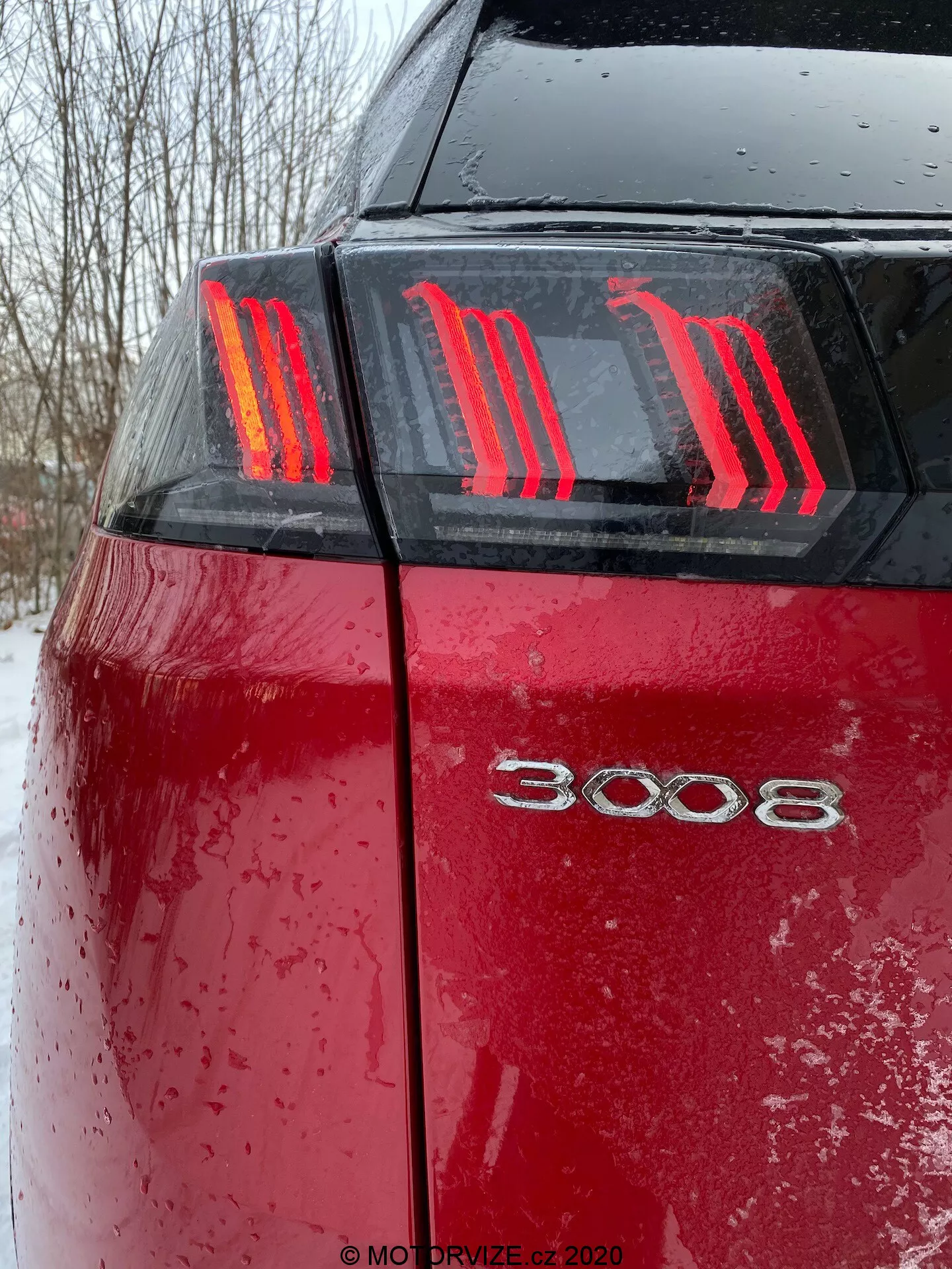 Detailní pohled na zadní část červeného Peugeot 3008 2021 z nízkého úhlu, ukazující výrazná tři vertikální LED světla vytvářející drápový vzor a modelové označení '3008'. Mokrá povrchová úprava auta, pokrytá kapkami vody a nečistotami nebo sněhem, zdůrazňuje nedávné použití v nepříznivém počasí s tlumenými světelnými podmínkami.