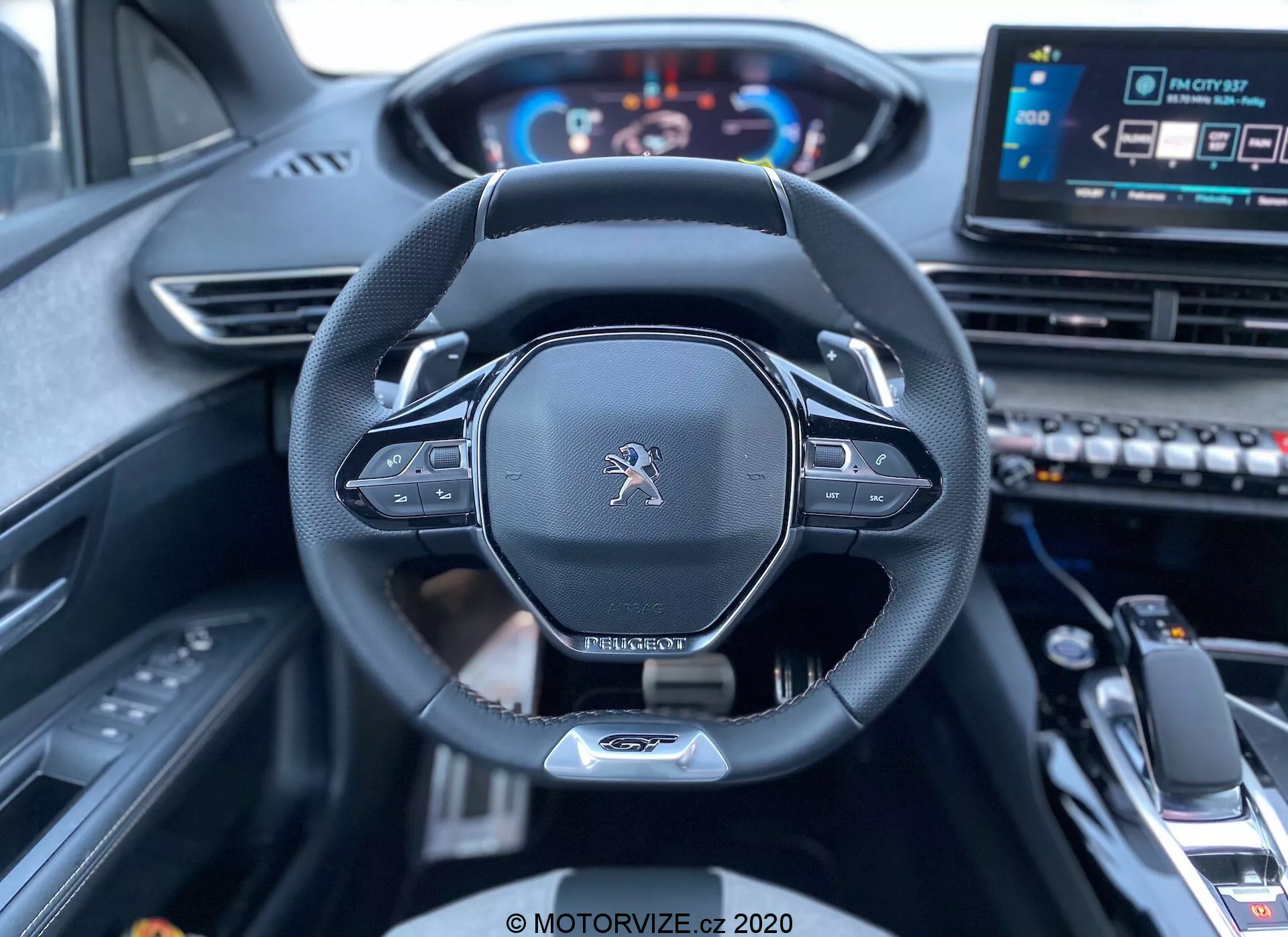 Pohled na interiér z řidičova sedadla vozu Peugeot 3008 z roku 2021, zobrazující volant a palubní desku. Snímek se zaměřuje na centrálně rámovaný volant, který má logo Peugeot, moderní design s plochým spodkem, kožený okraj a zabudované ovladače pravděpodobně pro nastavení audia a tempomatu. Za volantem jsou vidět páčky pro manuální řazení. Digitální přístrojový panel zobrazující rychloměr a otáčkoměr je osvětlený, což signalizuje, že je vozidlo zapnuté. Částečně viditelný je centrální dotykový infotainment systém a různé ovládací tlačítka vpravo, spolu s řadicí pákou a středovou opěrkou v dolní popředí, což naznačuje moderní, technologicky vyspělý vůz s luxusním interiérem.