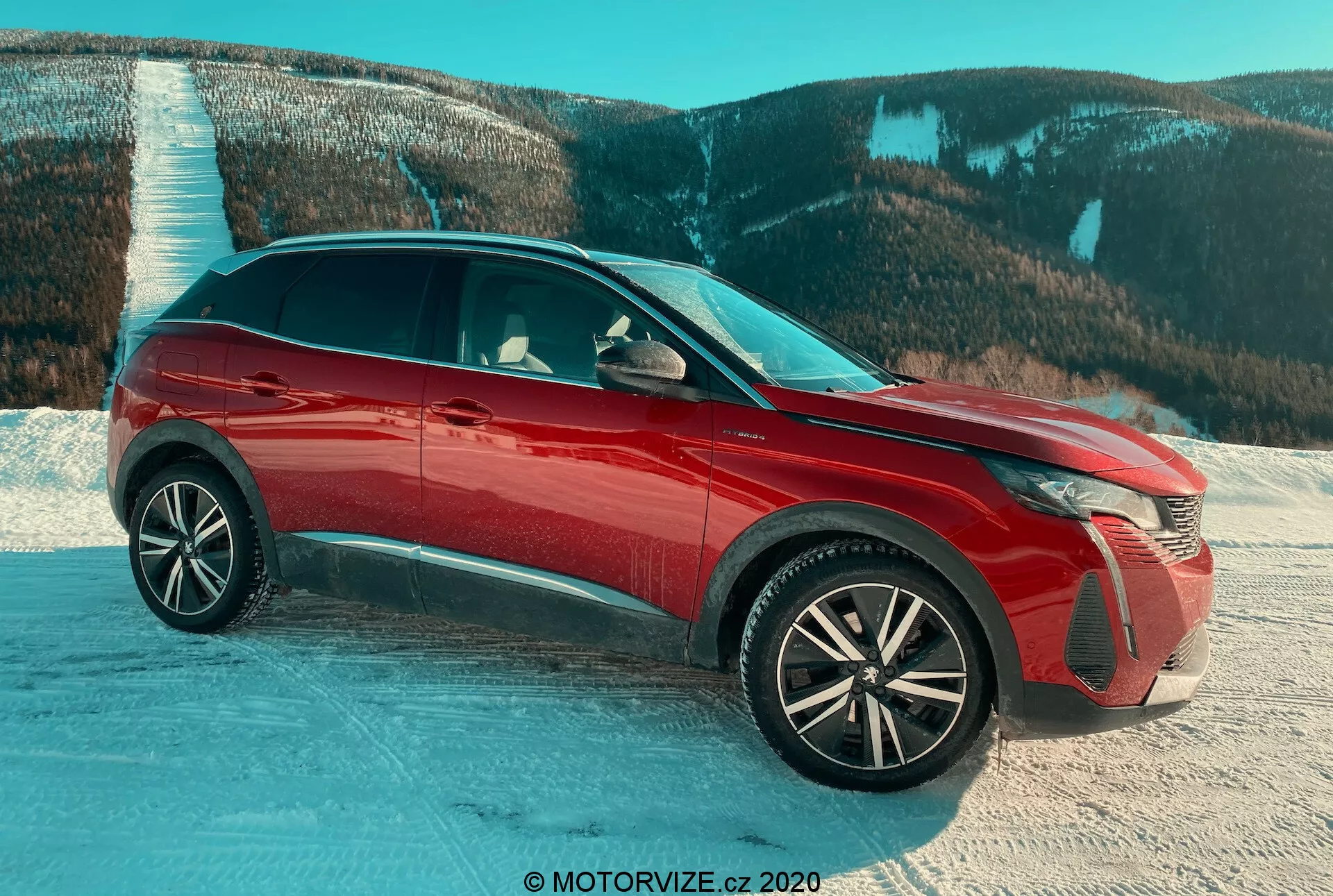 SUV Peugeot 3008 z roku 2021 v červené barvě, fotografováno z pohledu tři čtvrtiny zepředu na zasněženém povrchu s horskou krajinou v pozadí, s důrazem na pokročilé LED světlomety, propracovaná hliníková kola, lesklý povrch, aktivní rámečky chladiče a boční zrcátka s integrovanými ukazateli směru, zdůrazňující designové prvky a vhodnost pro náročné podmínky.