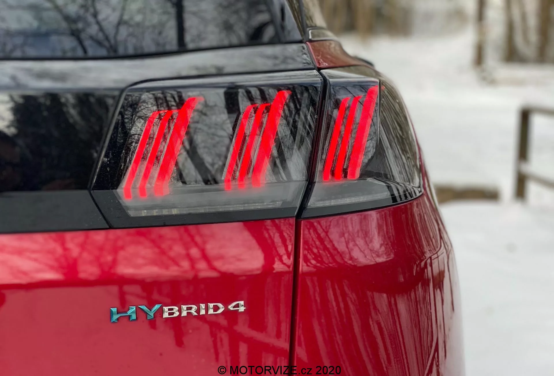 Peugeot 3008 Hybrid4 2021 - Detailní pohled na zadní levý čtvrtý úhel s živou červenou barvou, moderními červenými LED světly s vertikálními segmenty, víkem kufru a odznakem hybridu na zadním levém rohu, což naznačuje hybridní pohon s možnou pohonem všech kol.