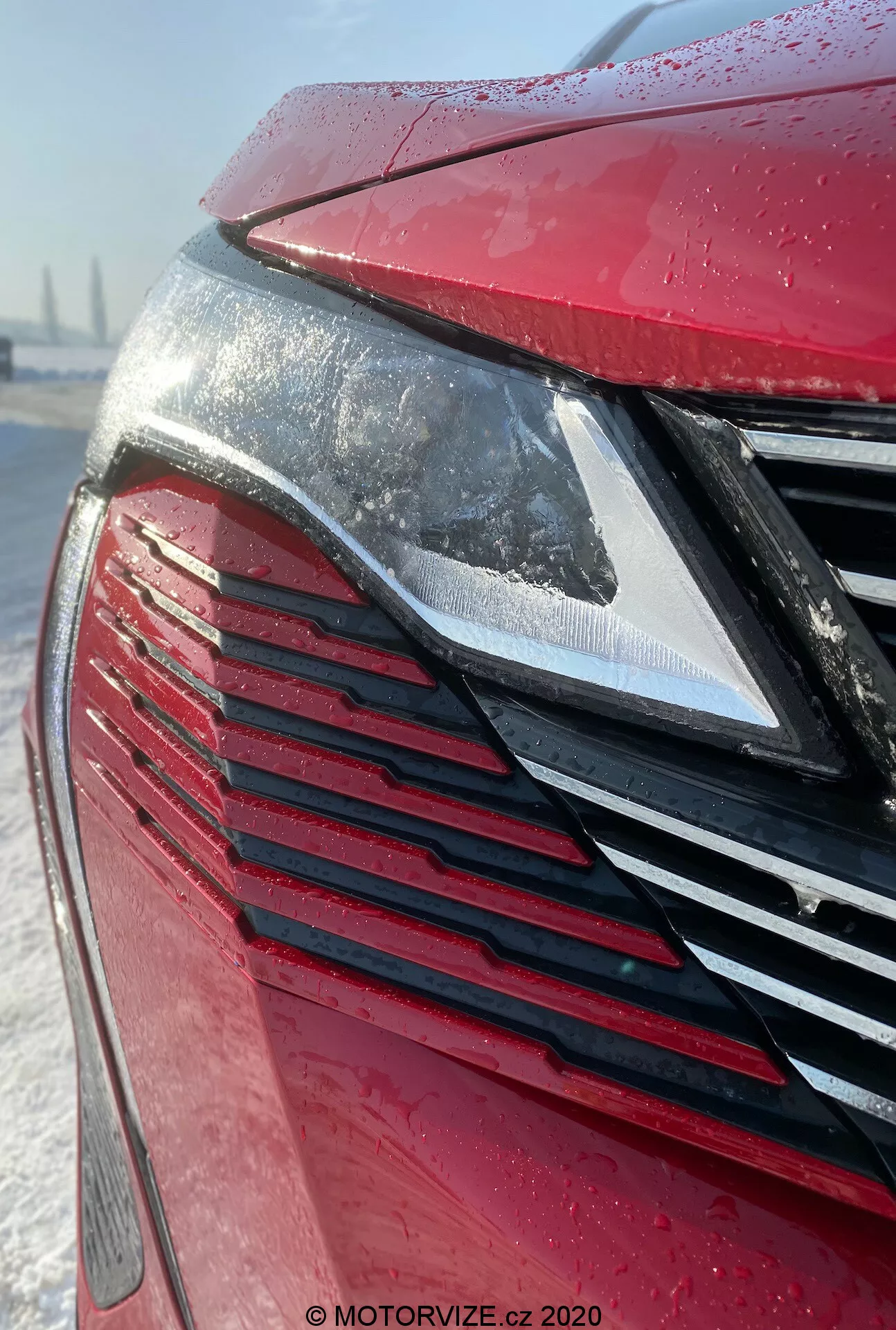 Detailní pohled na přední část vozu Peugeot 3008 2021 se zaměřením na moderní LED světlomet a mřížku s vertikálními lamelami v metalické červené barvě pokryté kapkami vody, což naznačuje nedávný déšť nebo jinovatku. Sněhové pozadí naznačuje zimní podmínky nebo péči o auto.