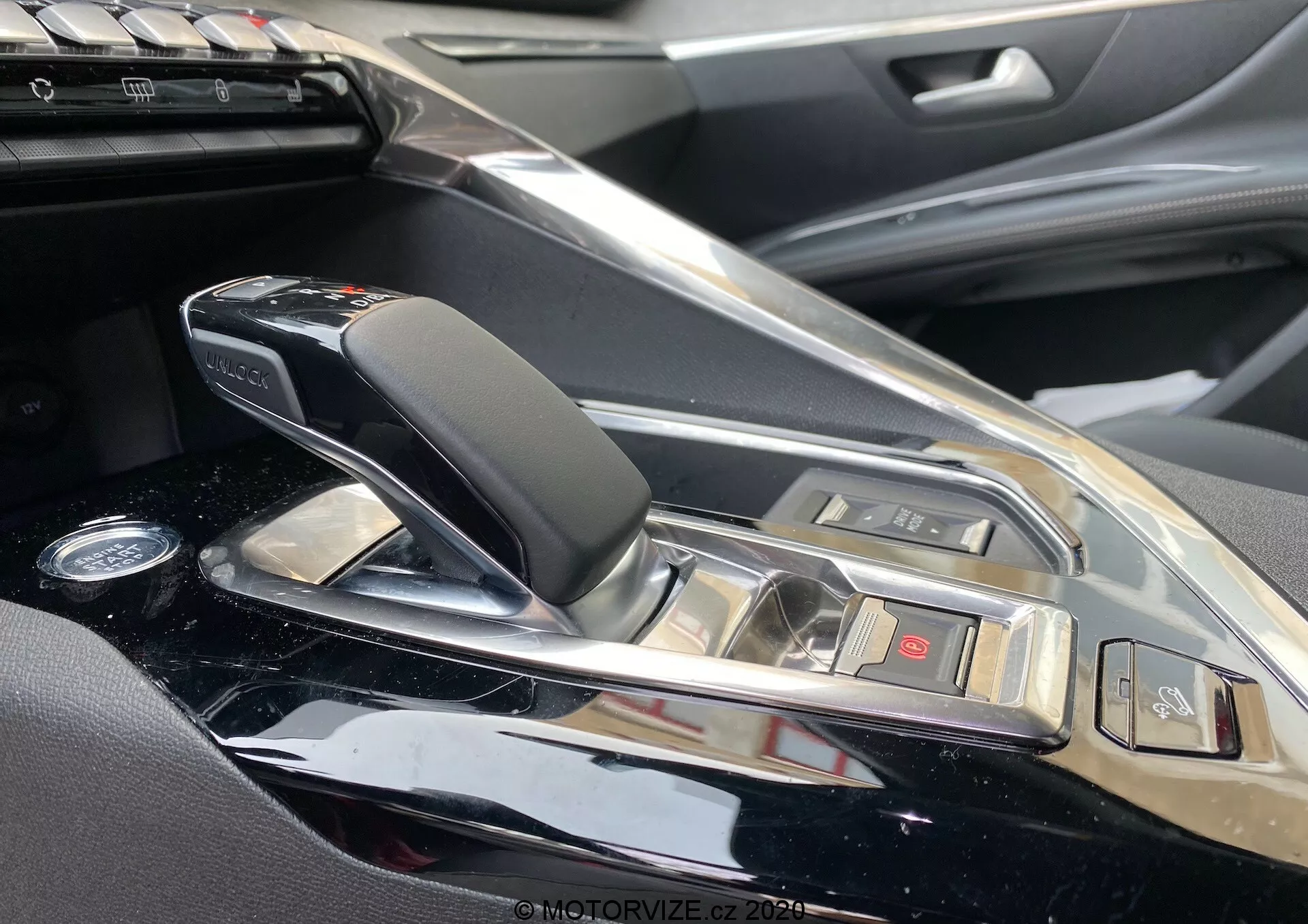 Detailní pohled na středovou konzolu a oblast řadicí páky Peugeotu 3008 2021 z pohledu spolujezdce. Obsahuje elegantní, moderní automatickou řadicí páku s koženými a chromovanými prvky, obklopenou vysoce lesklým povrchem konzoly. Viditelné jsou tlačítka pro různé funkce vozidla a náznak informačního systému, což naznačuje luxusní interiér vozidla s vysoce kvalitními materiály a sofistikovanými technologiemi.
