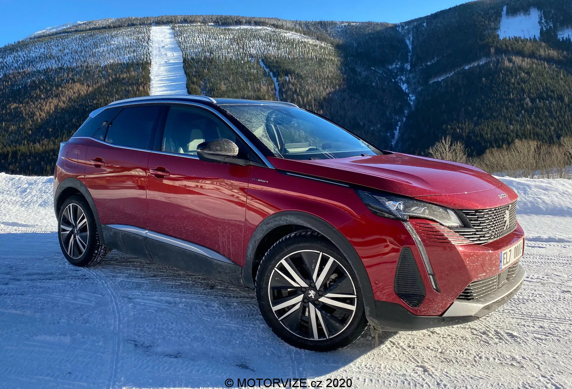 Obrázek moderního vozu, pravděpodobně Peugeot 3008 z roku 2021, pořízený z předního tříčtvrťového pohledu. Úhel ukazuje výrazný design LED světlometů vozu, mřížku s hřebenovým vzorem a znak na přední straně. Přední nárazník zobrazuje sekce sání vzduchu a pouzdra mlhových světel. Kapota má tvarované linie, které přispívají k aerodynamice, zatímco rozsáhlé čelní sklo naznačuje dobrou viditelnost vpřed. Kola z několika paprsků naznačují sportovnější a vyšší úroveň výbavy. Elegantní linie střechy a dynamické linie těla naznačují crossover nebo SUV. Viditelné jsou také boční zrcátka s integrovanými ukazateli směru a bočním lištováním nebo akcenty. Scéna je zasněžená silnice s horským pozadím, což naznačuje schopnost vozidla pro různé terény.