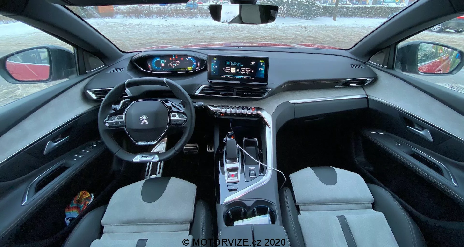 Pohled na interiér vozu Peugeot 3008 2021, ukazující multifunkční kožený volant s namontovanými ovládacími prvky, digitální přístrojový panel, velký dotykový infotainment systém na středové konzoli, elektronický volič rychlostí, přední sedadla s dvojitou barvou a směs praktických a luxusních materiálů. Záběr je pořízen ze zadního sedadla, poskytující rozsáhlý pohled na kabinu s důrazem na design a technologické funkce.