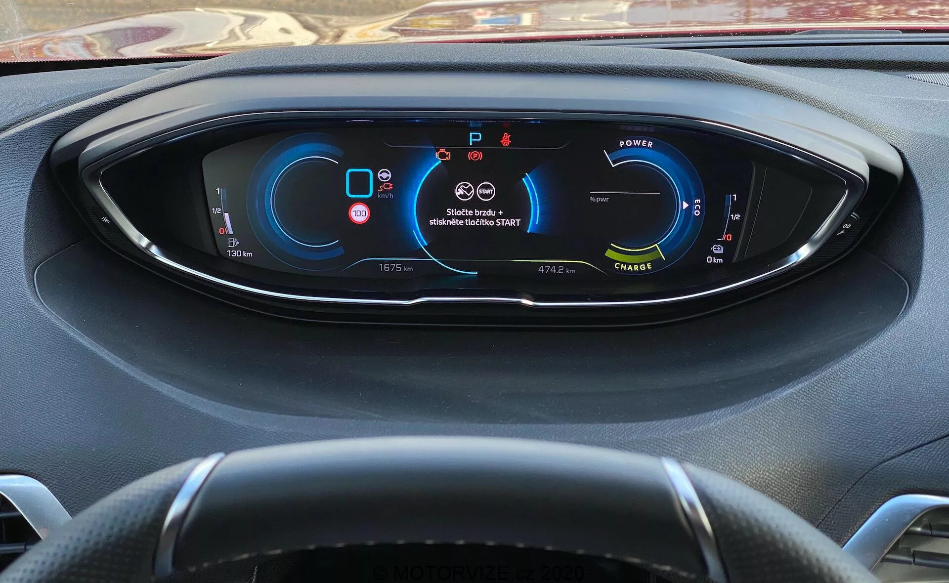 Pohled řidiče na panel přístrojů Peugeot 3008 z roku 2021 s plně digitálním displejem zobrazujícím metriky a indikátory vozidla. Vlevo je ukazatel výkonu naznačující elektrické/hybridní vozidlo; uprostřed nahoře jsou výstrahy pásu a airbagu; vpravo je tachometr ukazující stacionární rychlost; dole jsou údaje o rychlostních stupních (parkování), tachometru, venkovní teplotě, jízdních režimech a dostupném dojezdu. Částečně viditelný tříramenný volant. Pohled zpoza volantu s mírným sklonem dolů.