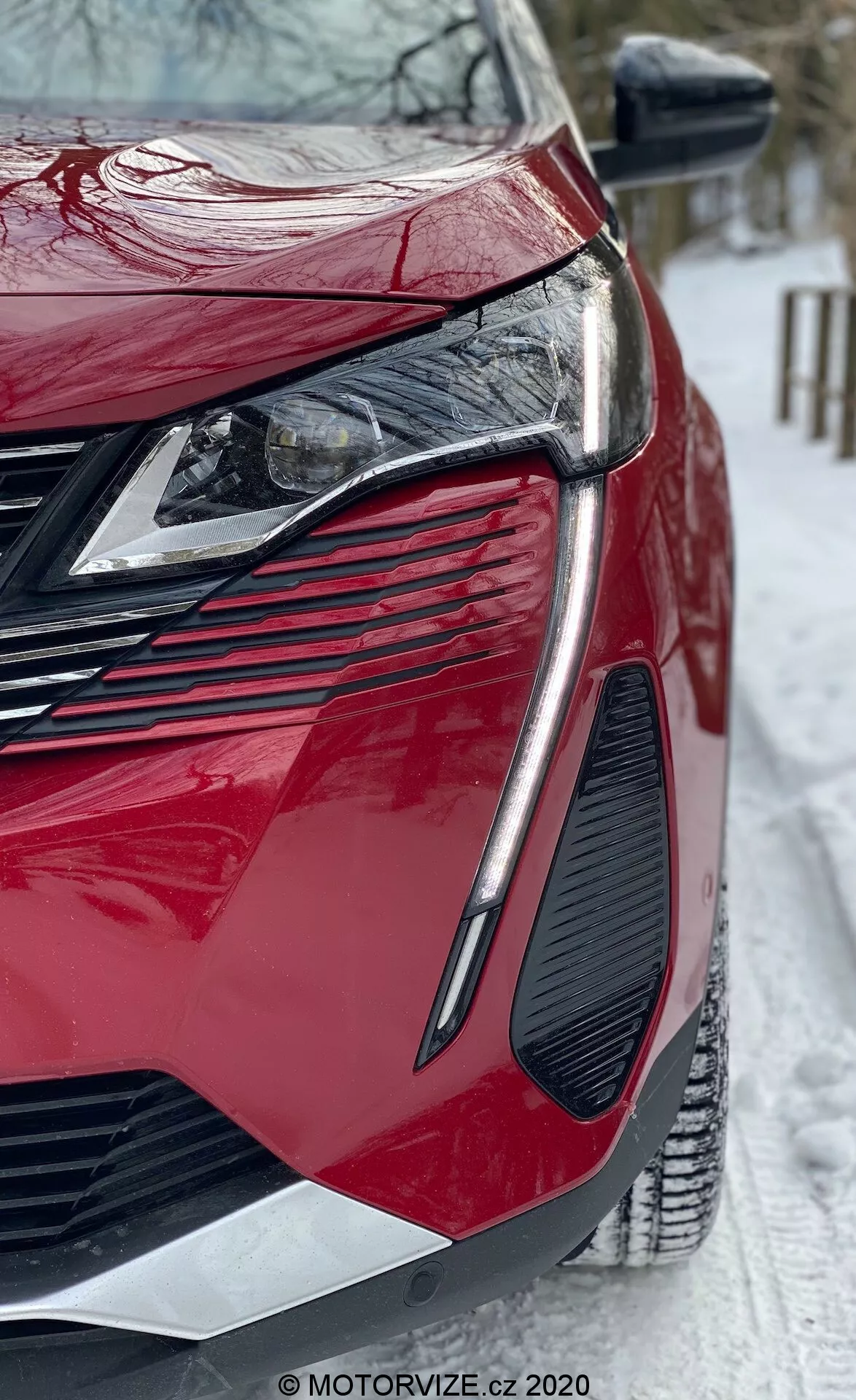 Detailní záběr z nízkého úhlu na moderní červený Peugeot 3008 2021. Zaměřeno na přední levý čtvrtletí, ukazující elegantní hranaté LED světlomety, velkou agresivní mřížku, výrazný design ventů, lesklý lak a ostře konturované karosářské panely. Zrcátko má integrované blinkry a scéna zahrnuje zasněženou zem, což naznačuje zimní podmínky. Viditelný vodoznak z MOTORVIZE.cz.