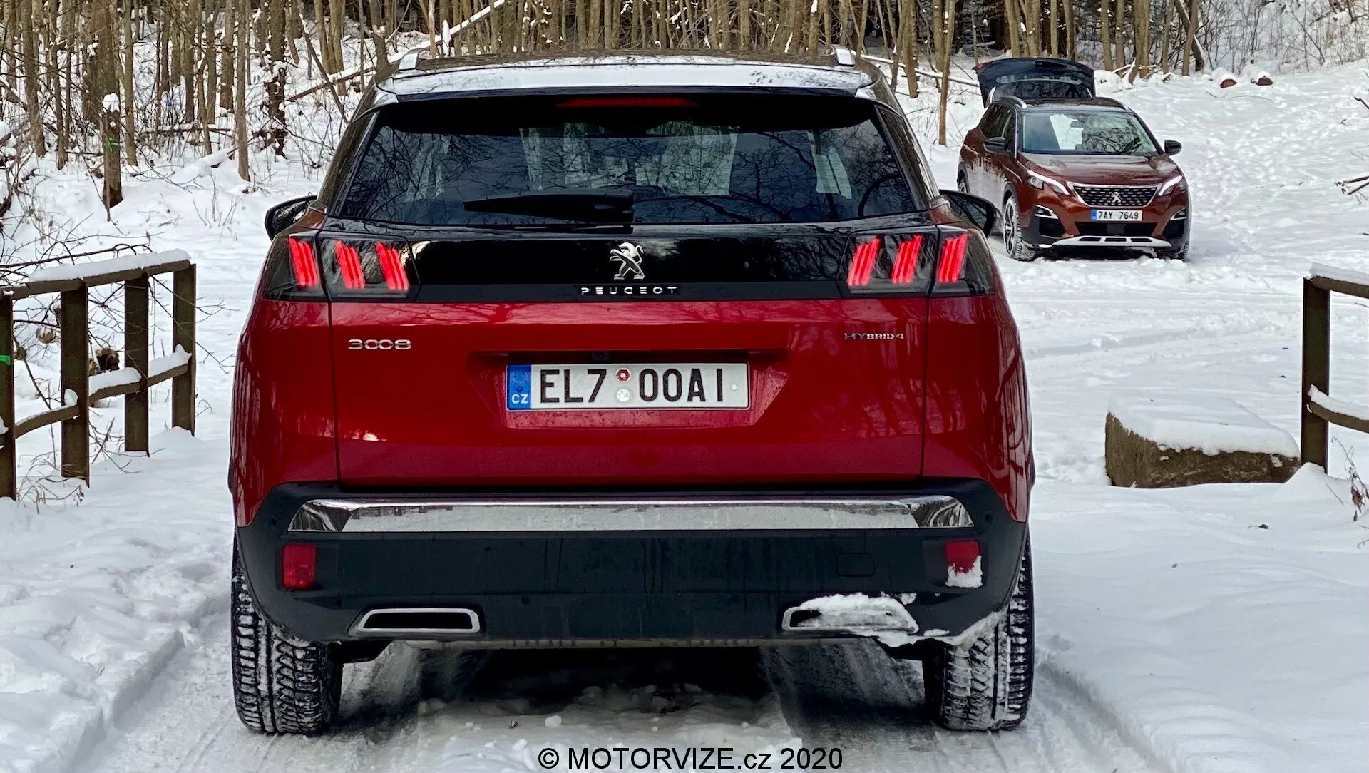 Zadní pohled na červený Peugeot 3008 SUV zachycený z mírného nízkého úhlu, zdůrazňující jeho charakteristická třípruhová spárová zadní světla, centrální znak Peugeot Lion a dvoutónový nárazník. Vozidlo, umístěné na sněhem pokryté zemi, naznačuje zimní podmínky. Viditelné funkce zahrnují zadní parkovací senzory, stěrač zadního okna a anténu v zadní části nahoře, naznačující technologii asistence řidiče a potenciální GPS/radio funkčnost, vhodné pro diskusi o automobilovém designu, bezpečnostních funkcích a zimním řízení.