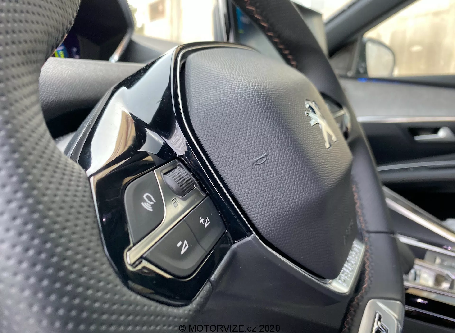 Detailní pohled na volant z pohledu řidiče v modelu Peugeot 3008 2021. Obrázek zdůrazňuje levou stranu volantu, pořízený z mírně nízkého úhlu a zachycující podrobnosti ovládacích prvků na levé straně a logo Peugeot. Volant je pokryt černou perforovanou kůží s kombinací lesklých a matných černých materiálů. Viditelné ovládací prvky zahrnují přepínač, tlačítka pro mediální a komunikační funkce jako přijímání nebo ukončení hovorů, aktivaci hlasového ovládání a další neidentifikovanou funkci. Ostrost je vysoká, ukazuje prošívání na kůži a lesklý povrch některých plastových částí, možná dotyková tlačítka.
