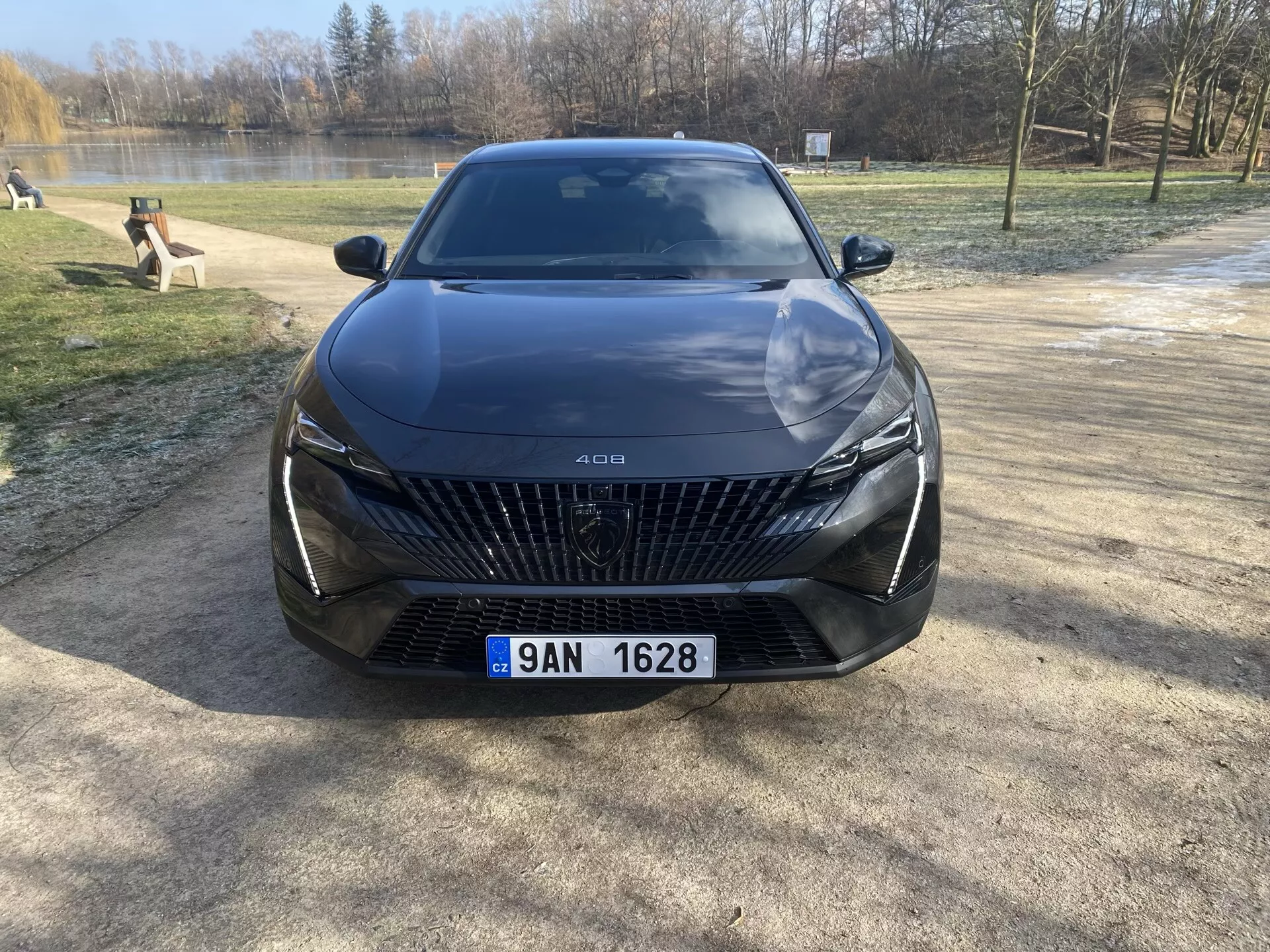Přední pohled na Peugeot 408 2023 zaparkovaný na štěrkovém povrchu s pozadím stromů a vody, zdůrazňující jeho velkou mřížku s diamantovými vzory, elegantní LED světlomety, sochařsky pojaté linie kapoty a pokročilé funkce asistence řidiče