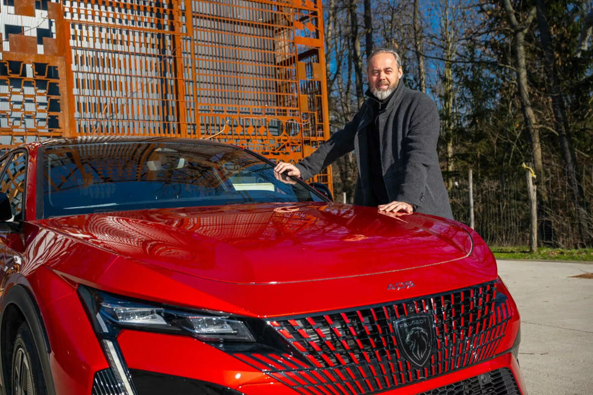 Červený model Peugeot 408 rok 2023 předvádějící svůj přední levý čtvrtletí ve dne, zdůrazňující detailní LED světlomety, geometrickou mřížku a sportovně aerodynamickou kapotu s mužem v tmavém kabátu stojícím vedle a pokládajícím si ruku na kapotu