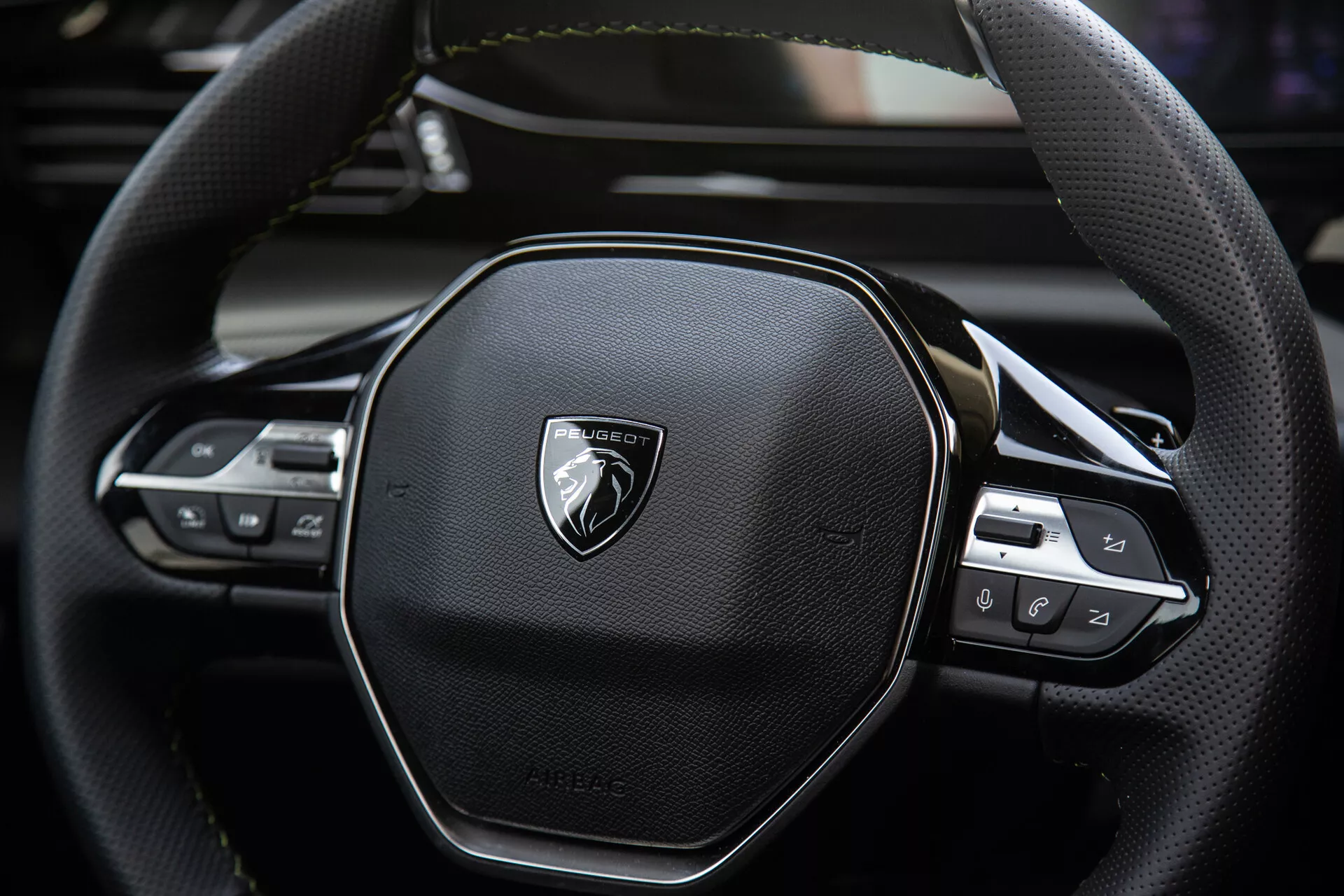 Detailní pohled na volant Peugeot 408 2023 s ergonomickým šestiúhelníkovým designem, prošíváním z kůže, integrovanými ovládacími prvky pro audio a nastavení tempomatu, s emblémem na kartáčovaném kovu.