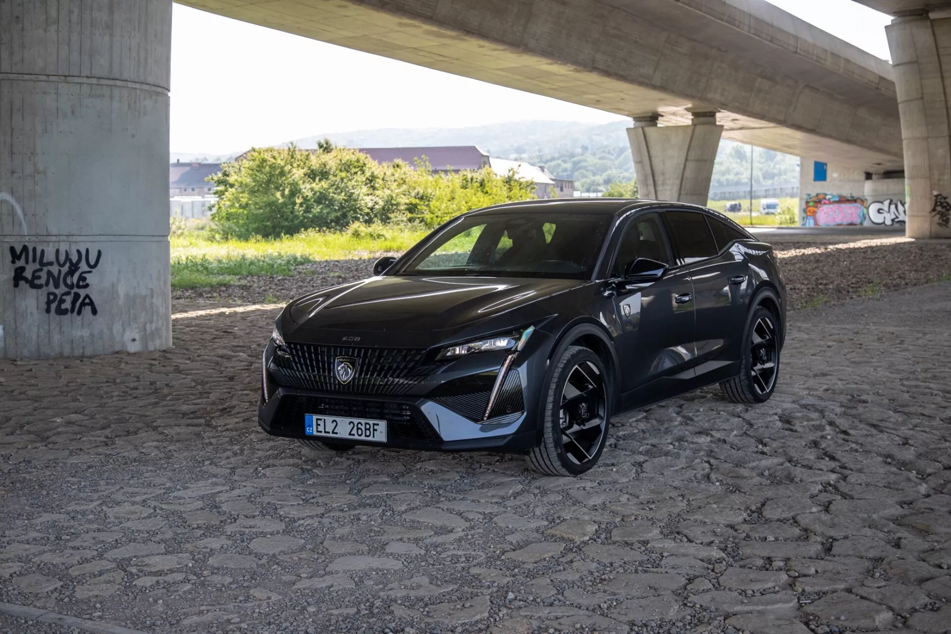 Model Peugeot 408 2023 zachycený z úhlu tři čtvrtiny zdůrazňující jeho hladká LED světlomety, vzorovaná mřížka a sportovní aerodynamický přední nárazník pod přirozeným denním světlem na zpevněné ploše pod nadjezdem