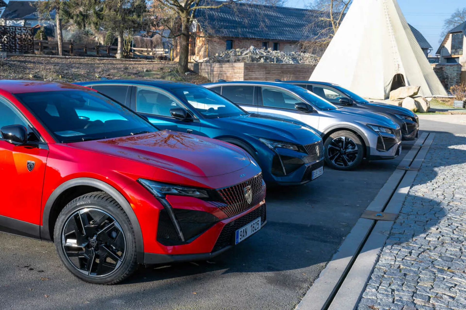 Řada vozů Peugeot 408 z roku 2023 s červeným vozem v popředí se sportovními vícepaprskovými koly a LED světlomety naznačujícími elektrický nebo hybridní model, zaparkovaná na dlážděné ploše vedle dlažby