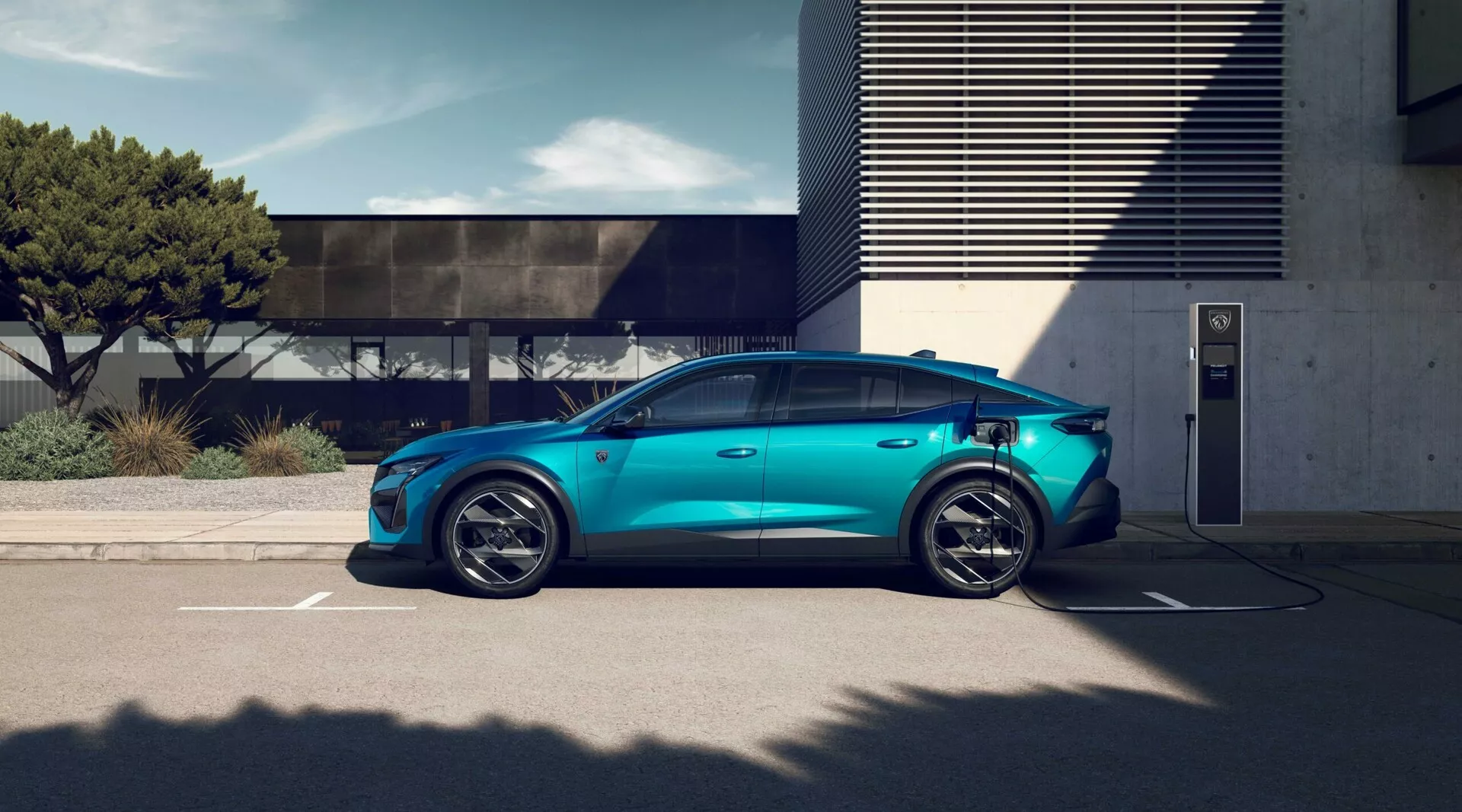 Elektrické vozidlo Peugeot 408 2023 v metalické modré barvě aparkované u venkovní dobíjecí stanice, předvádějící sportovně laděné pětipaprskové slitinové kola, aerodynamický siluetu, zapuštěné kliky dveří, viditelný dobíjecí port, konvenční boční zrcátka, okna s potemněním pro soukromí a panoramatickou střešní okno pod jasnými denními světelnými podmínkami s moderní architekturou na pozadí