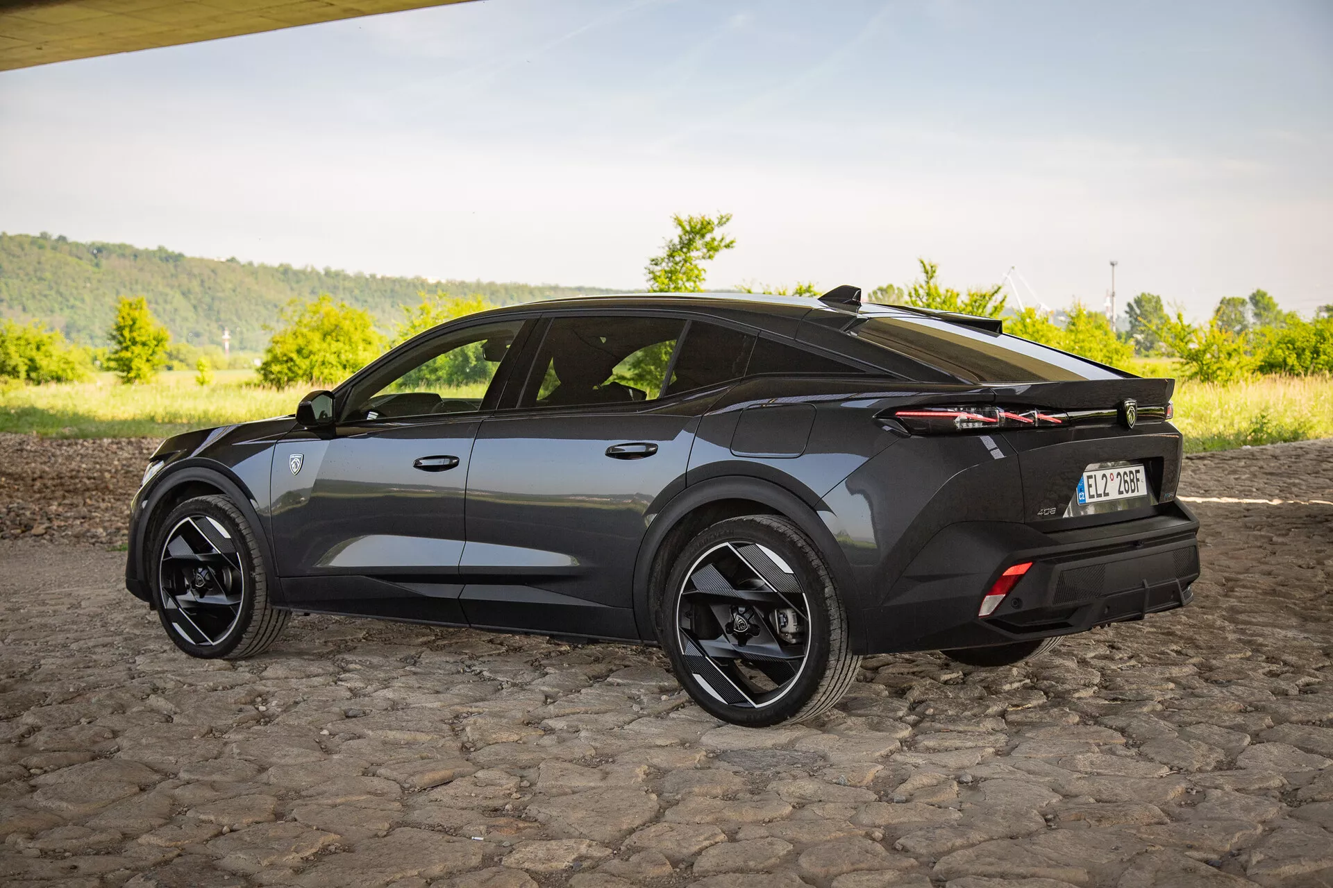 Crossover SUV Peugeot 408 roku 2023 ve tmavém provedení, se záběrem ze tříčtvrtečního zadního úhlu zdůrazňujícím aerodynamický design s ostrými úhly, sportovní skloněnou střechou, velkými černými slitinovými koly, výraznými LED zadními světly a decentním zadním spoilerem na dlážděném povrchu