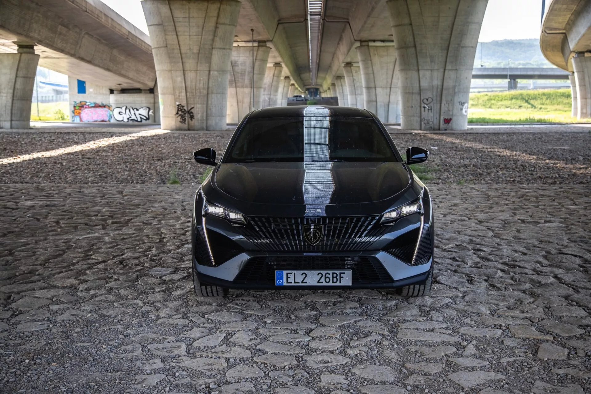 Peugeot 408 z roku 2023 v dynamickém pohledu zepředu zdůrazňující jeho agresivní postoj a sportovní design s detailní mřížkou, štíhlými LED světlomety a aerodynamickými prvky na vzorované dlažbě pod městským mostem