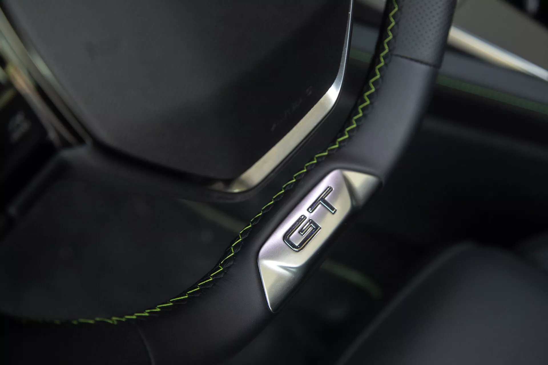 Detailní pohled na část volantu a palubní desky Peugeot 408 2023, černý kožený volant s zeleným prošíváním a odznakem GT, což zdůrazňuje luxus a sportovní charakter