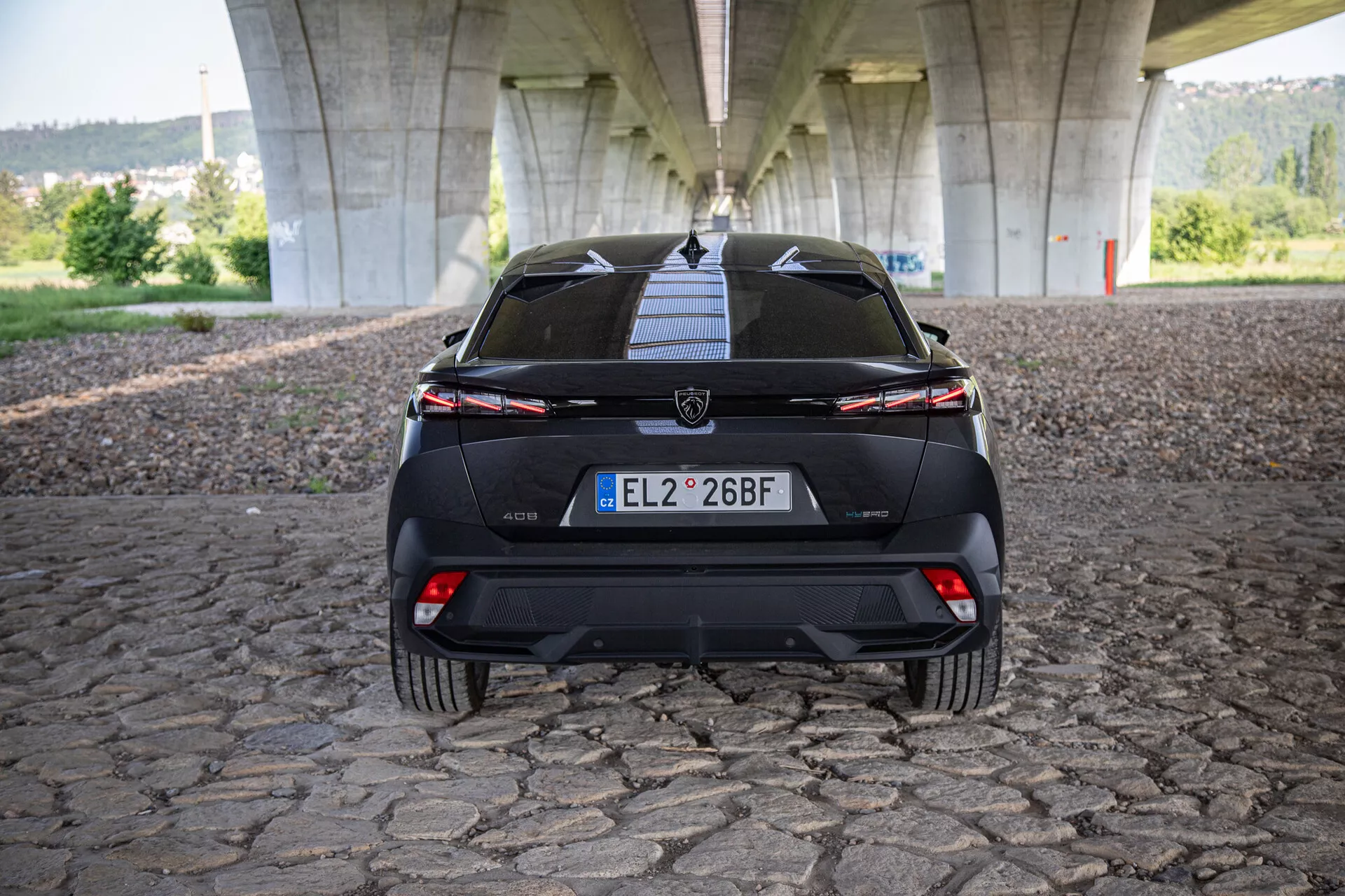 Zadní pohled na Peugeot 408 2023 zobrazující páté dveře, nárazník, spodní difuzor, zadní světla s připojujícím se světelným pruhem a senzory pro asistenci parkování, fotografováno pod mostem na dlažební ploše