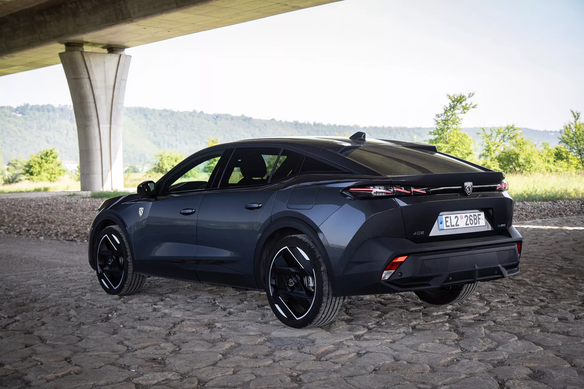 Tříčtvrteční zadní pohled na hatchback Peugeot 408 rok 2023 pod mostem na dlažebních kostkách se sportovními ráfky a pokročilou světelnou technologií