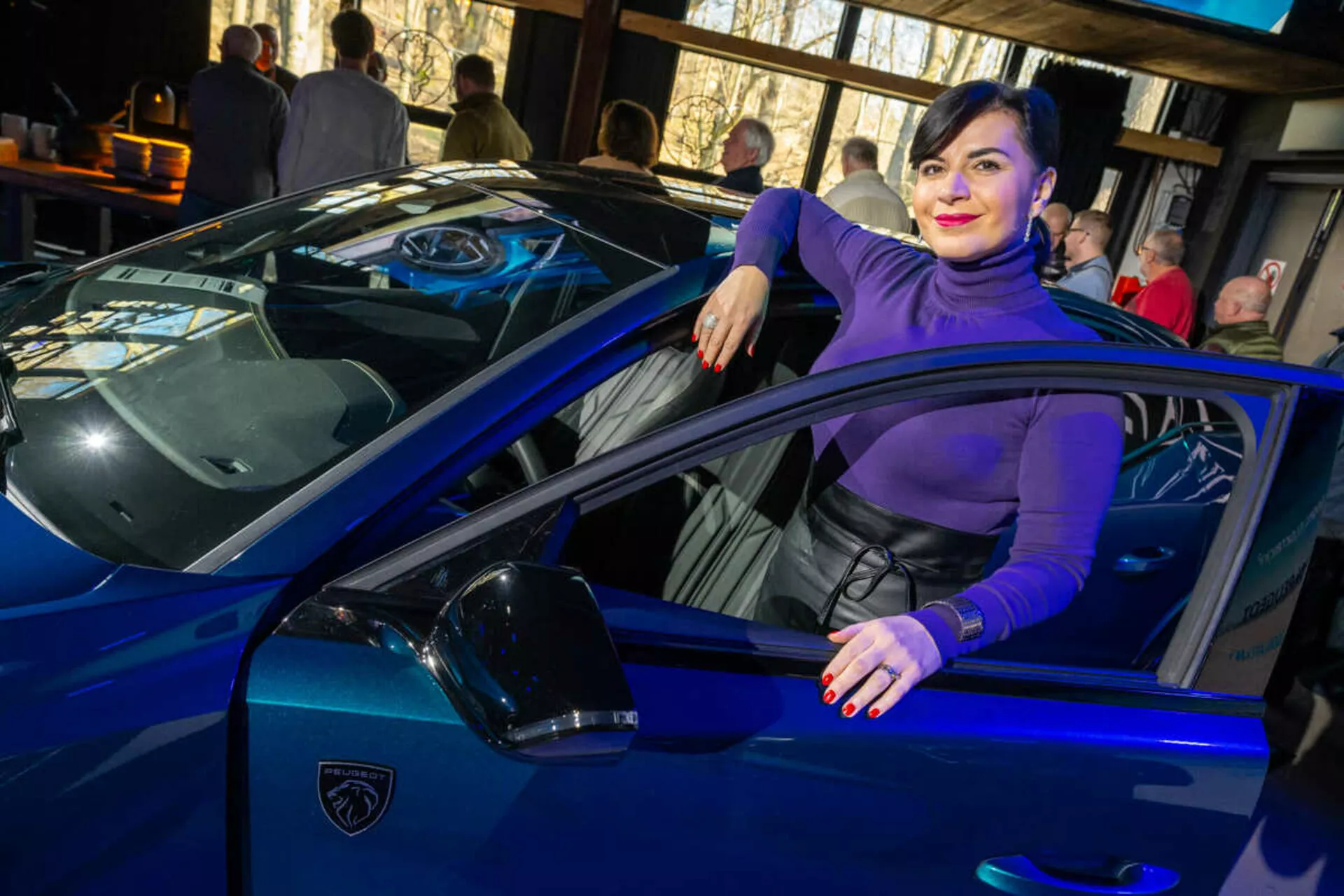 Živě modré sportovní auto, možná Lamborghini, zachycené ze tříčtvrtečního úhlu zdůrazňující jeho elegantní linie, ostré LED světlomety a ikonické štítové logo, s osobou neformálně interagující s vozidlem naznačující luxus a styl.
