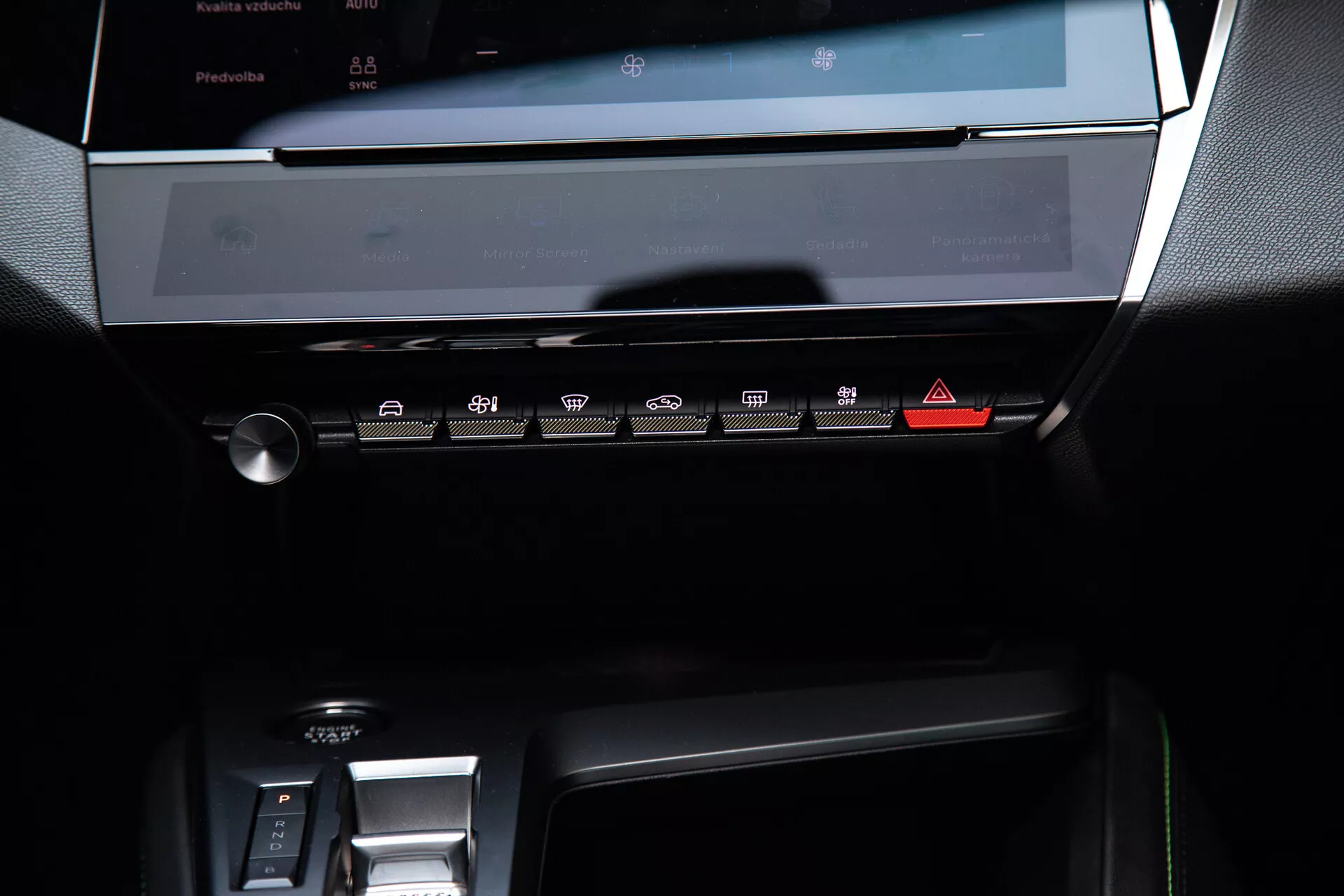 Centrální palubní deska Peugeotu 408 z roku 2023 s vypnutým dotykovým displejem infotainment systému, panelem klimatizace s dotykovými tlačítky a částí oblasti elektronického řazení za jemného ambientního osvětlení zdůrazňujícího její moderní, elegantní design s tmavými tóny a kovovými akcenty.